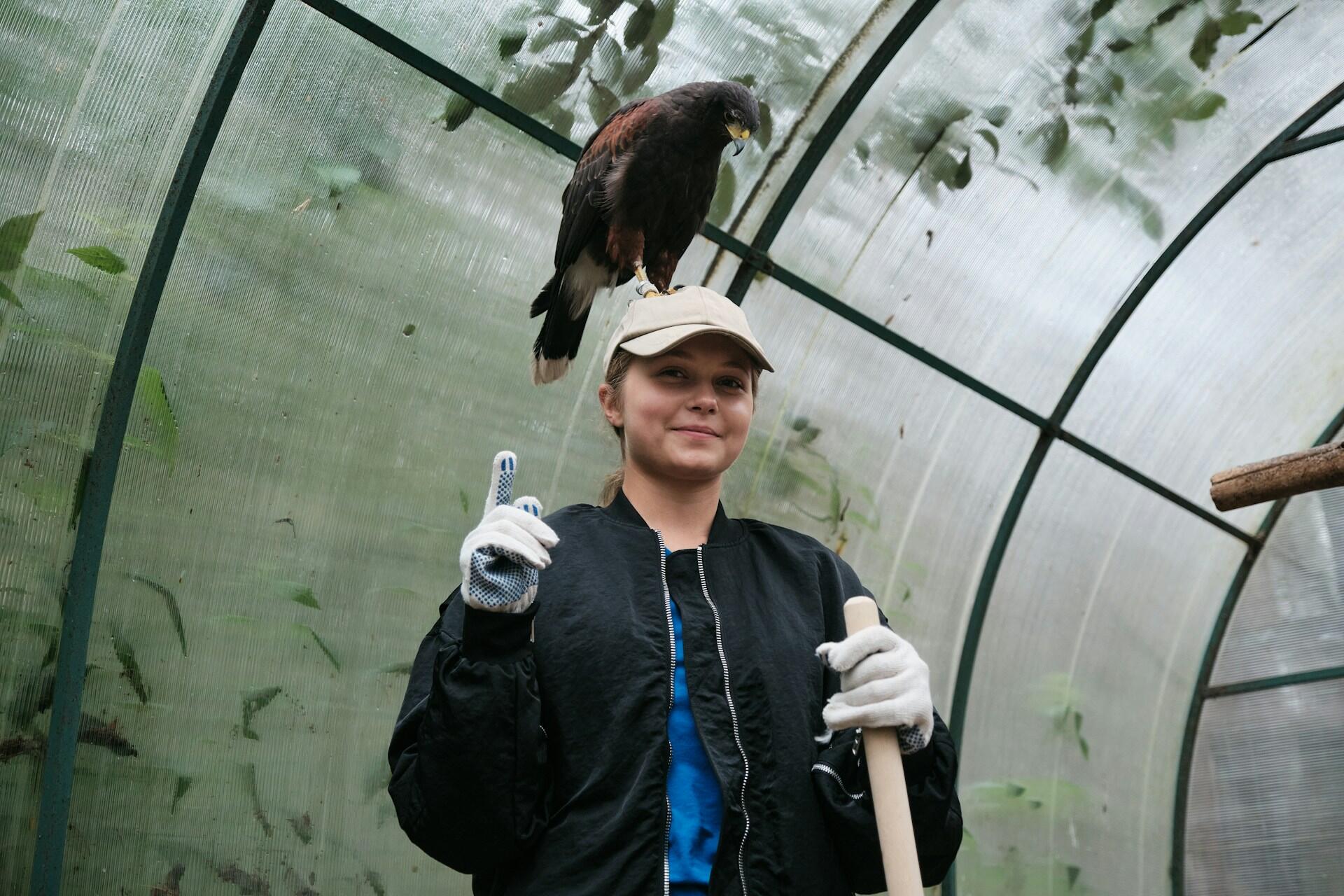 A woman in a greenhouse with a bird on her head.