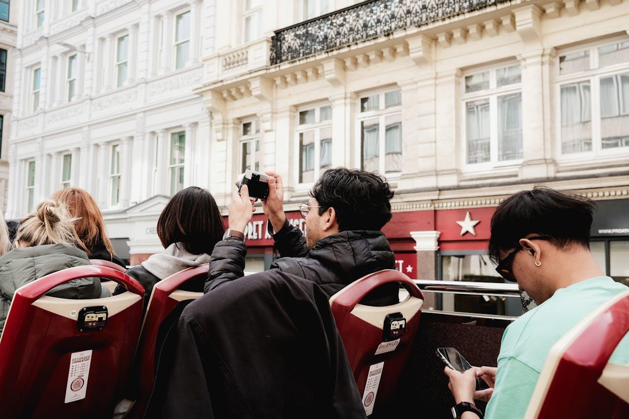 A group of people sitting on the top deck of a double-decker bus, engaged in taking photos and using their phones.