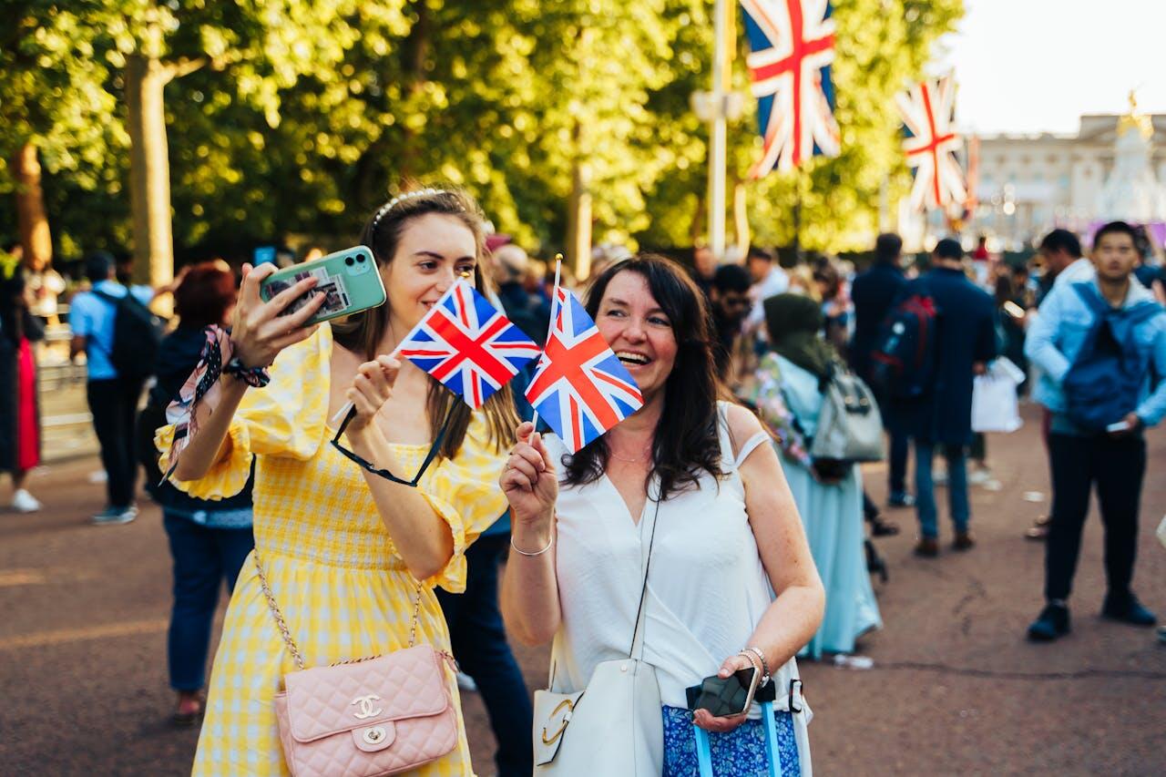 Two women celebrate with British flags in a lively crowd, surrounded by trees and Union Jack banners at a festive outdoor event.