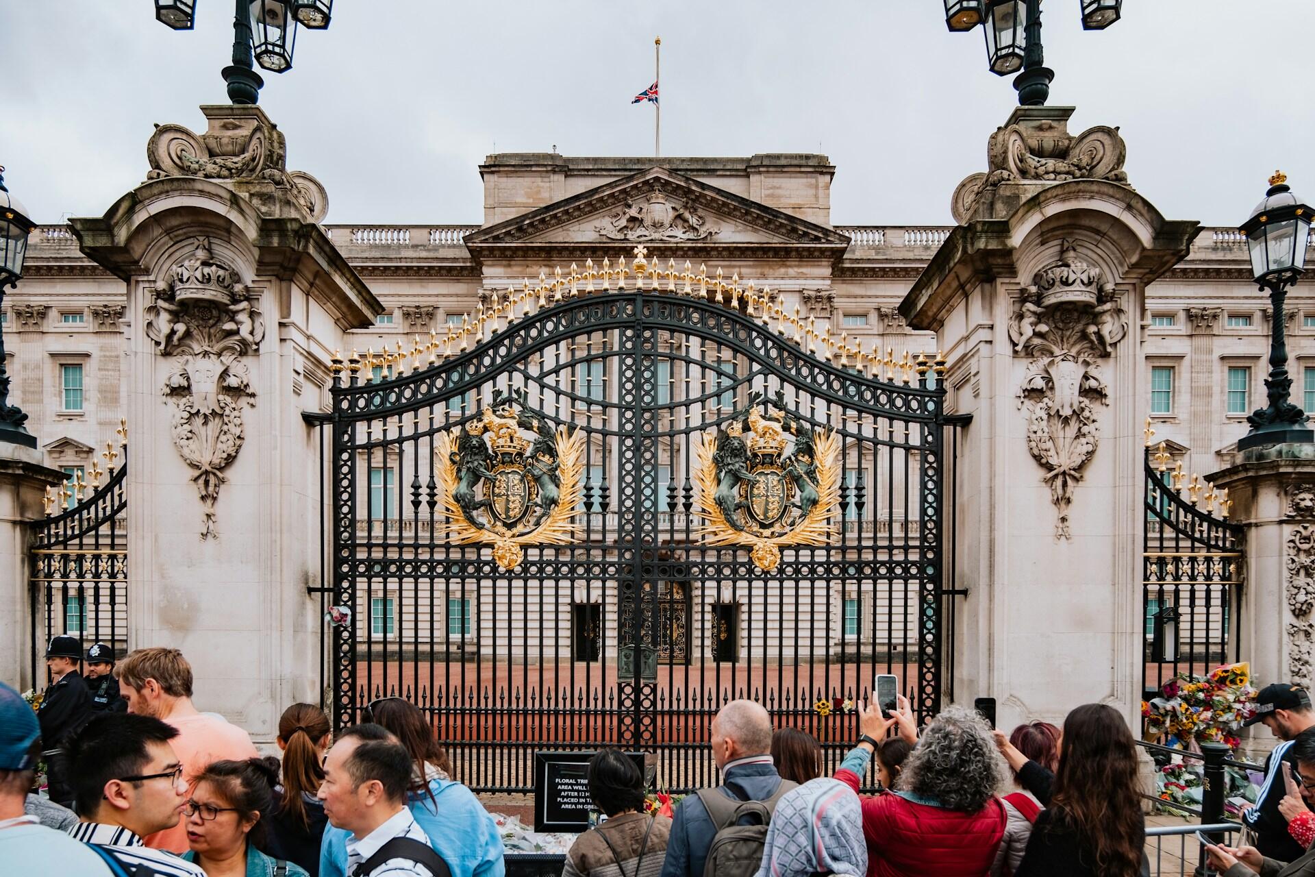 Visitors gather at the ornate gates of Buckingham Palace, with the iconic royal insignia prominently displayed.