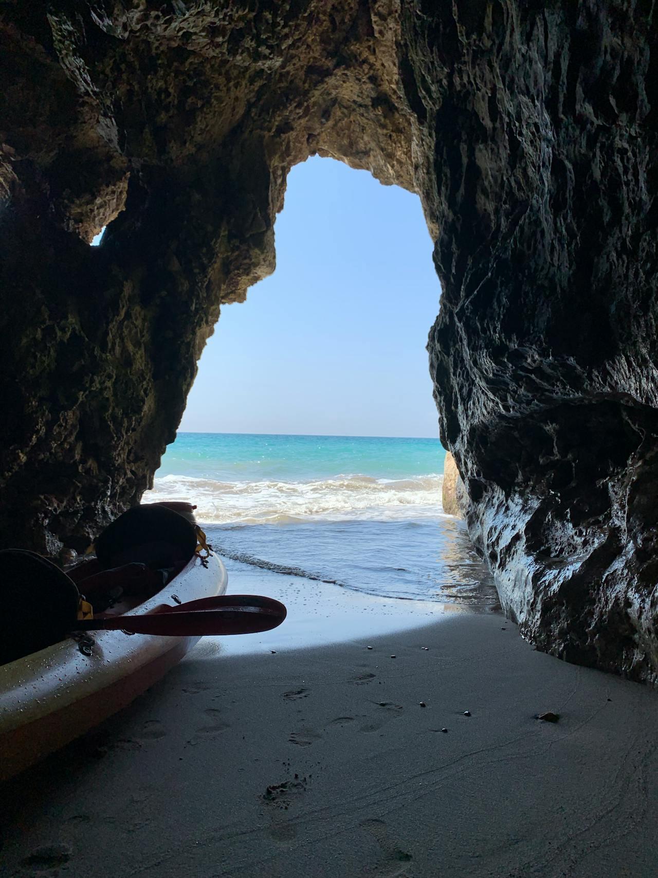 A kayak rests on sandy ground inside a rocky cave, framing a view of the blue ocean and sky beyond.
