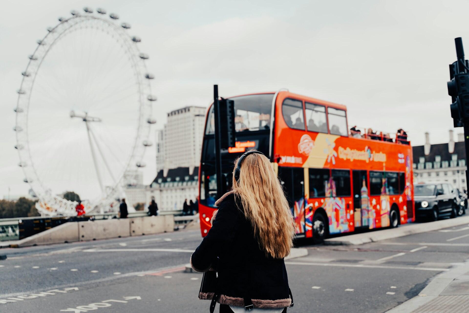 A woman with long hair stands on a street corner, watching a red double-decker bus near the London Eye in the background.