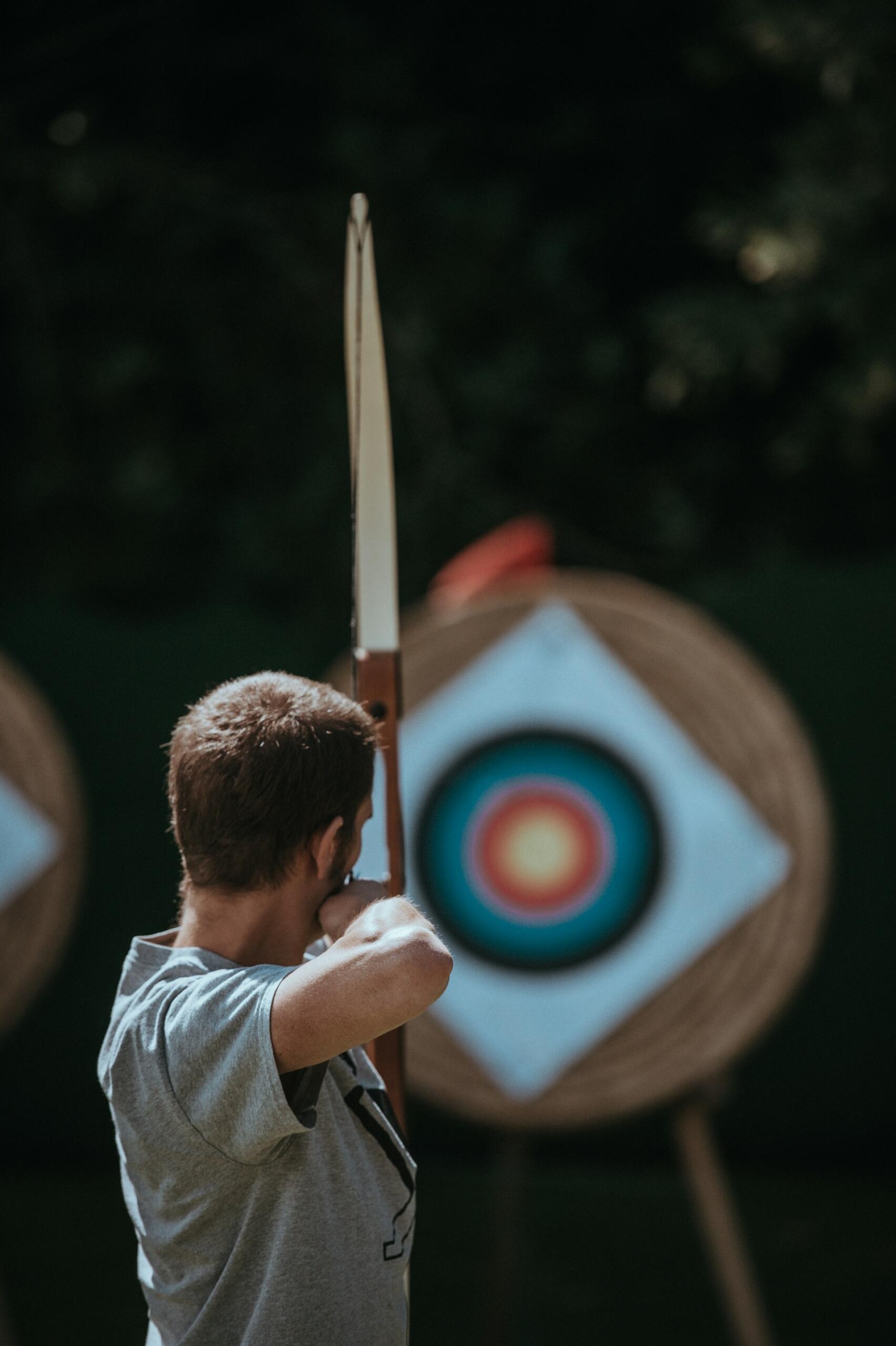 A person stands poised with a bow, aiming at a colorful archery target in the background, surrounded by greenery.
