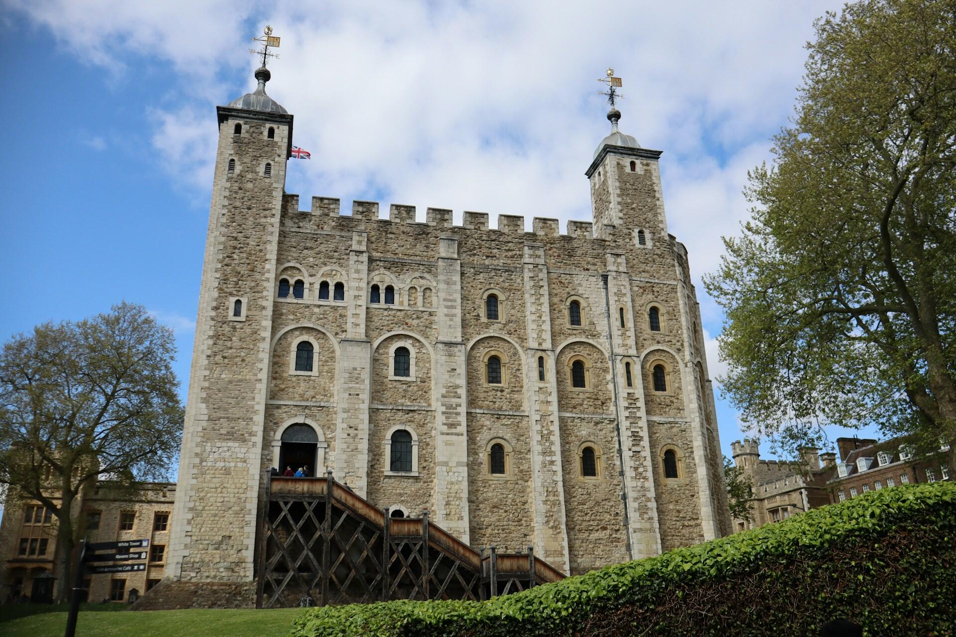 A historic stone castle with two towers, featuring arched windows, flags, and surrounded by green trees against a blue sky.