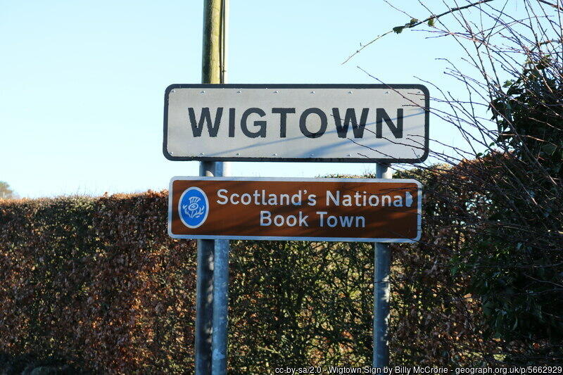 Road sign for Wigtown, Scotland, highlighting it as the National Book Town, surrounded by greenery and clear blue skies.