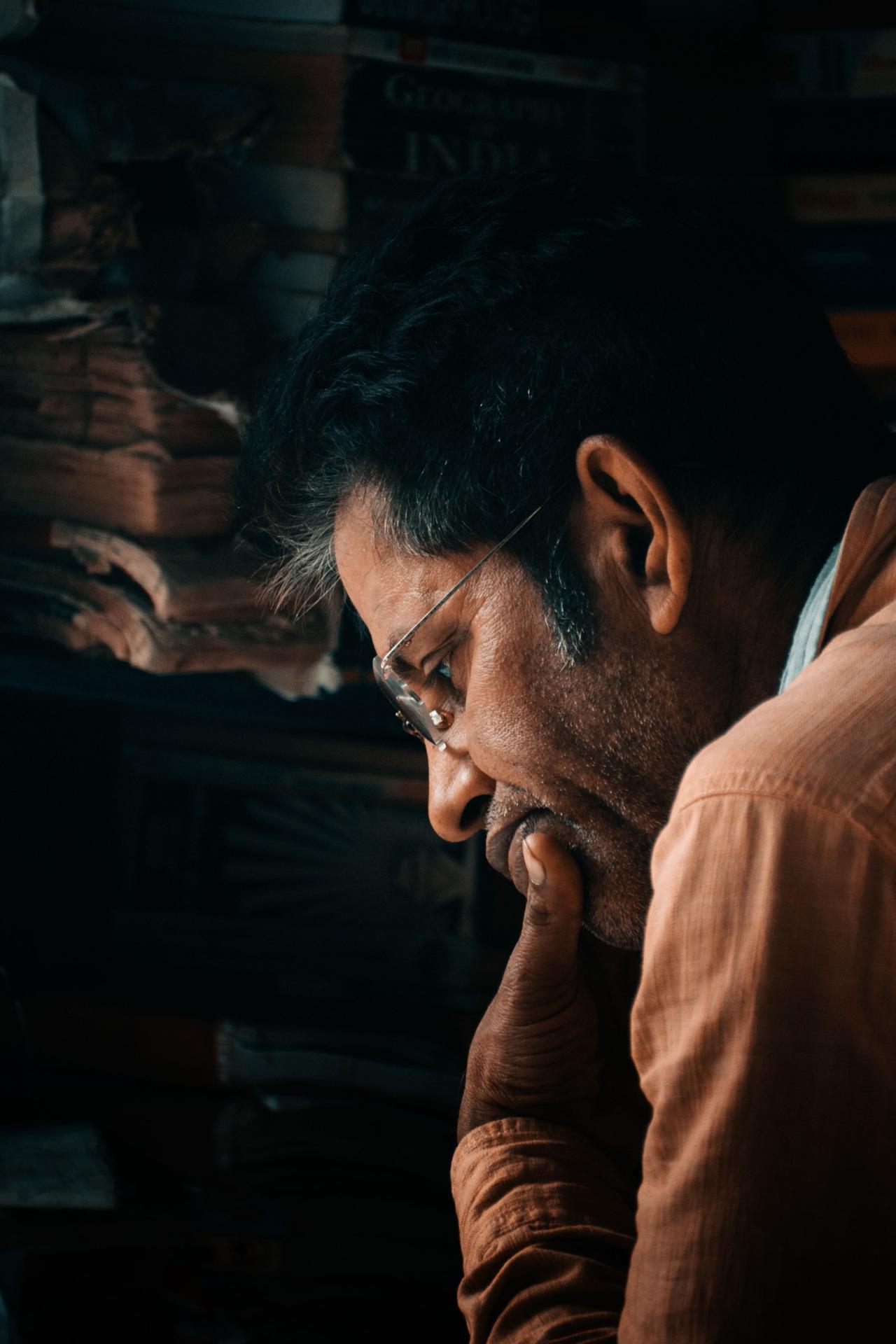 Man reading thoughtfully with books and newspapers around him, representing focused learning of Hindi verbs, grammar, and sentence structure.