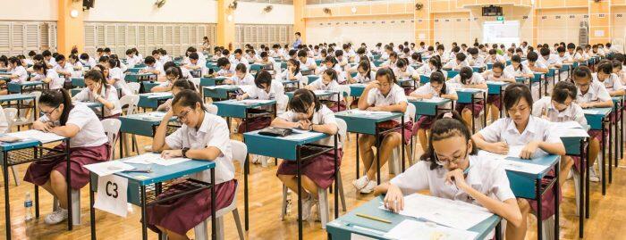 Students sit in long rows at desks, quietly writing exams in a bright school hall; the crowded scene feels focused and orderly.