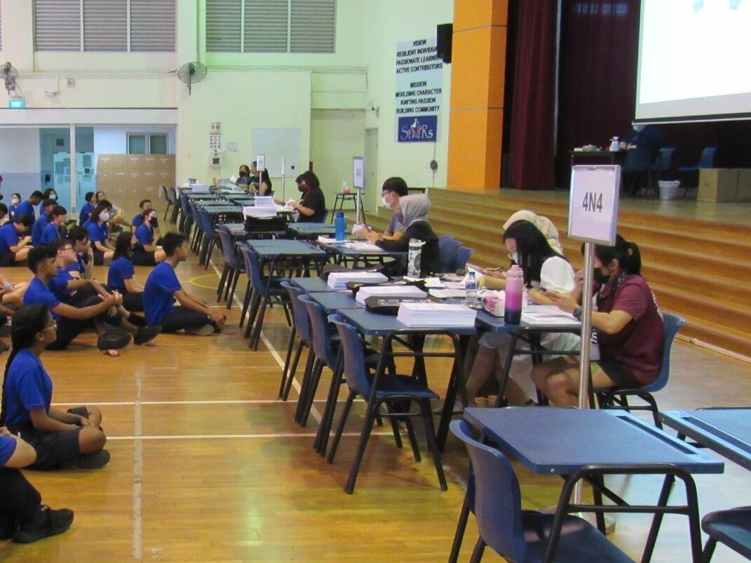 Students queue to register at tables in a school gym, while staff check forms; the busy, orderly scene feels focused and welcoming.