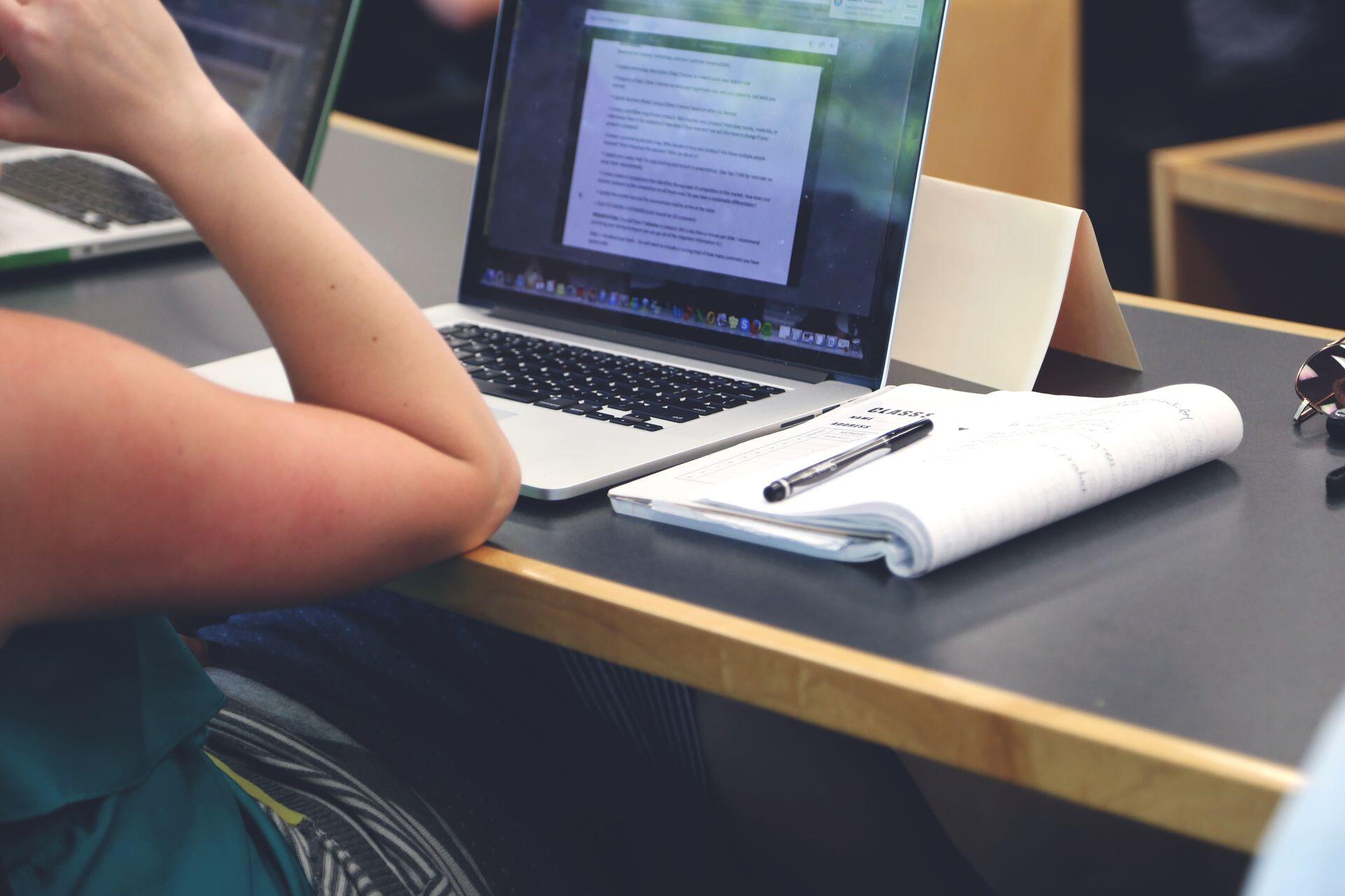 A student in a classroom setting using a laptop and taking notes in a notebook.