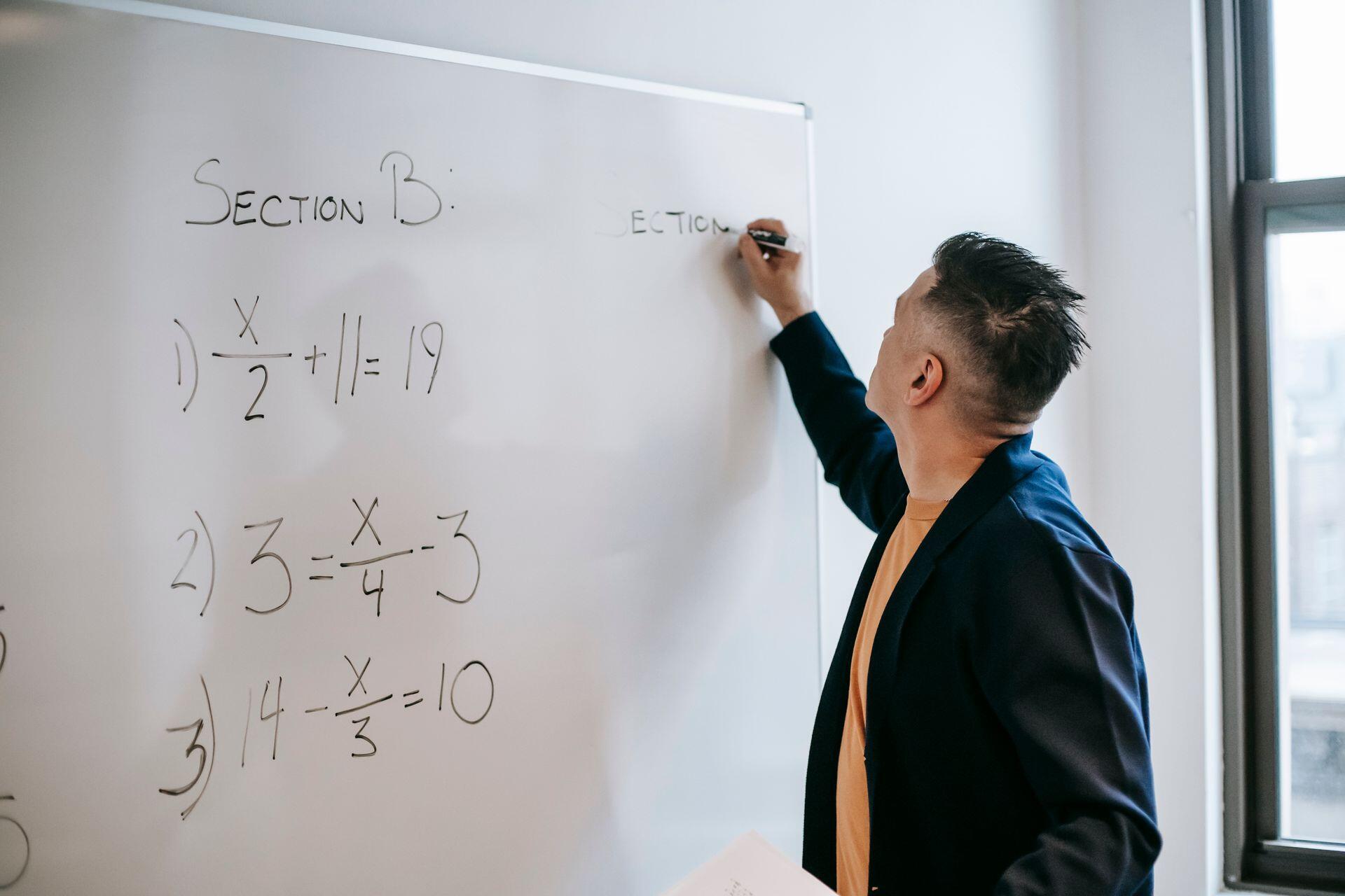 Teacher writing mathematical equations on a whiteboard in a classroom setting, focused on teaching.