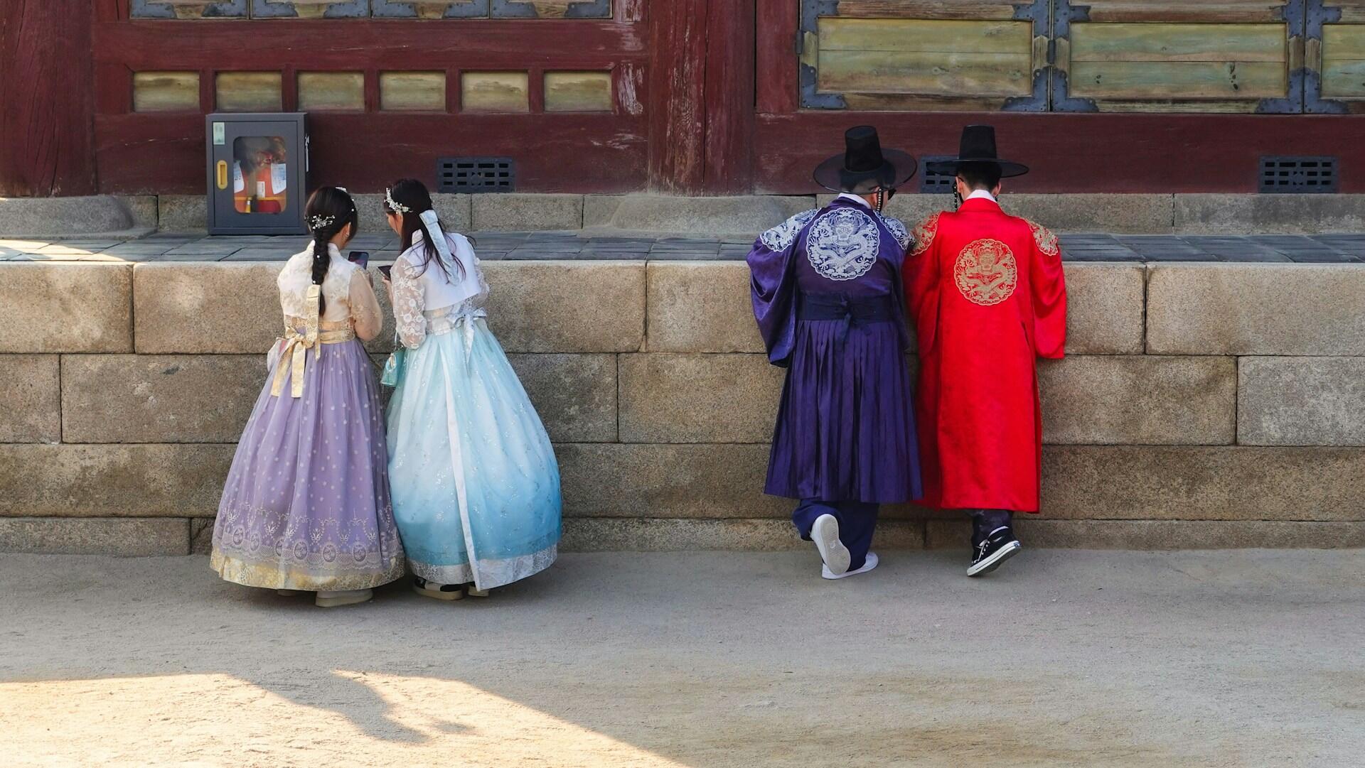 Four people wearing hanbok by a low stone wall.