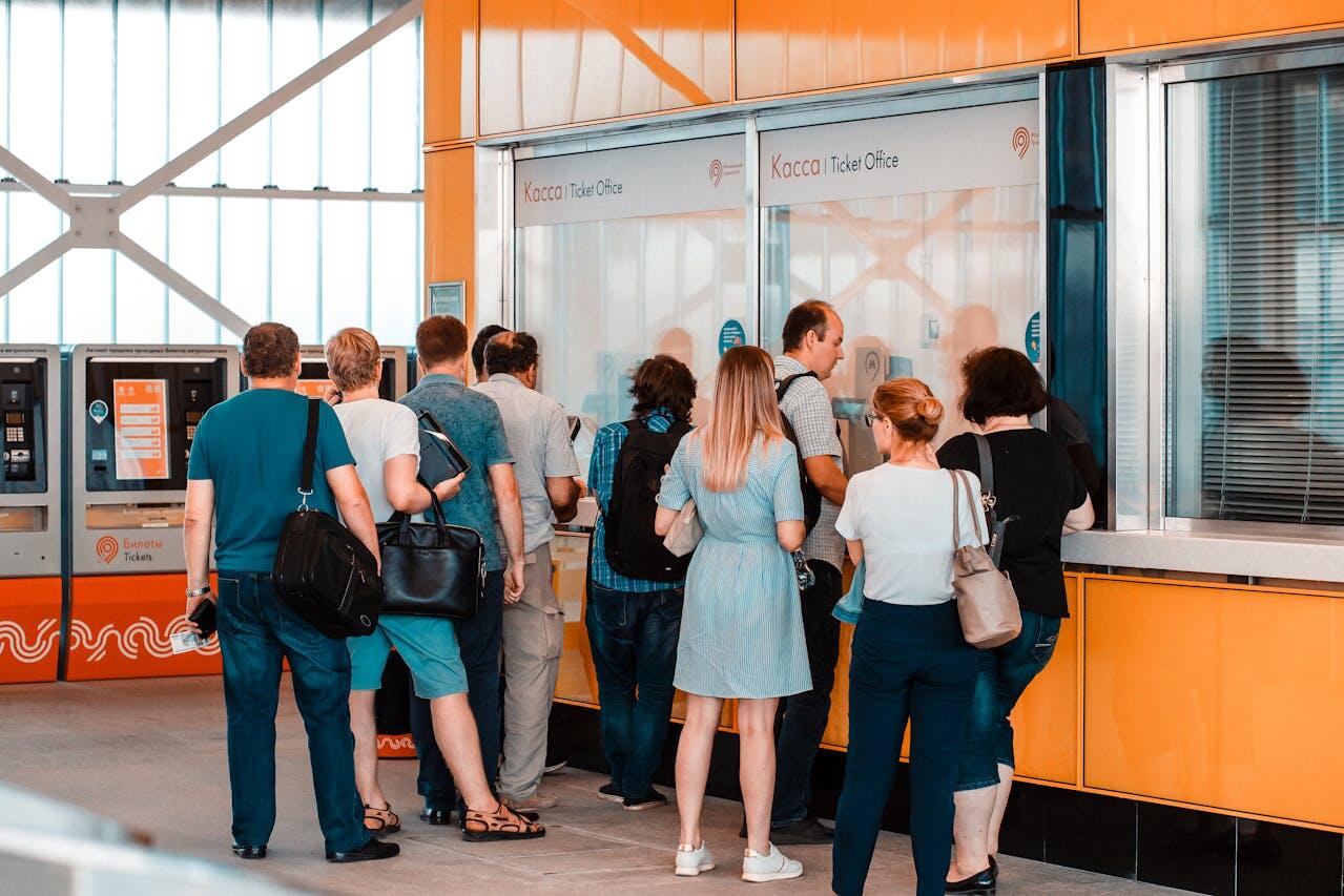 A group of people waiting in line at a brightly colored ticket office, with vending machines visible in the background.