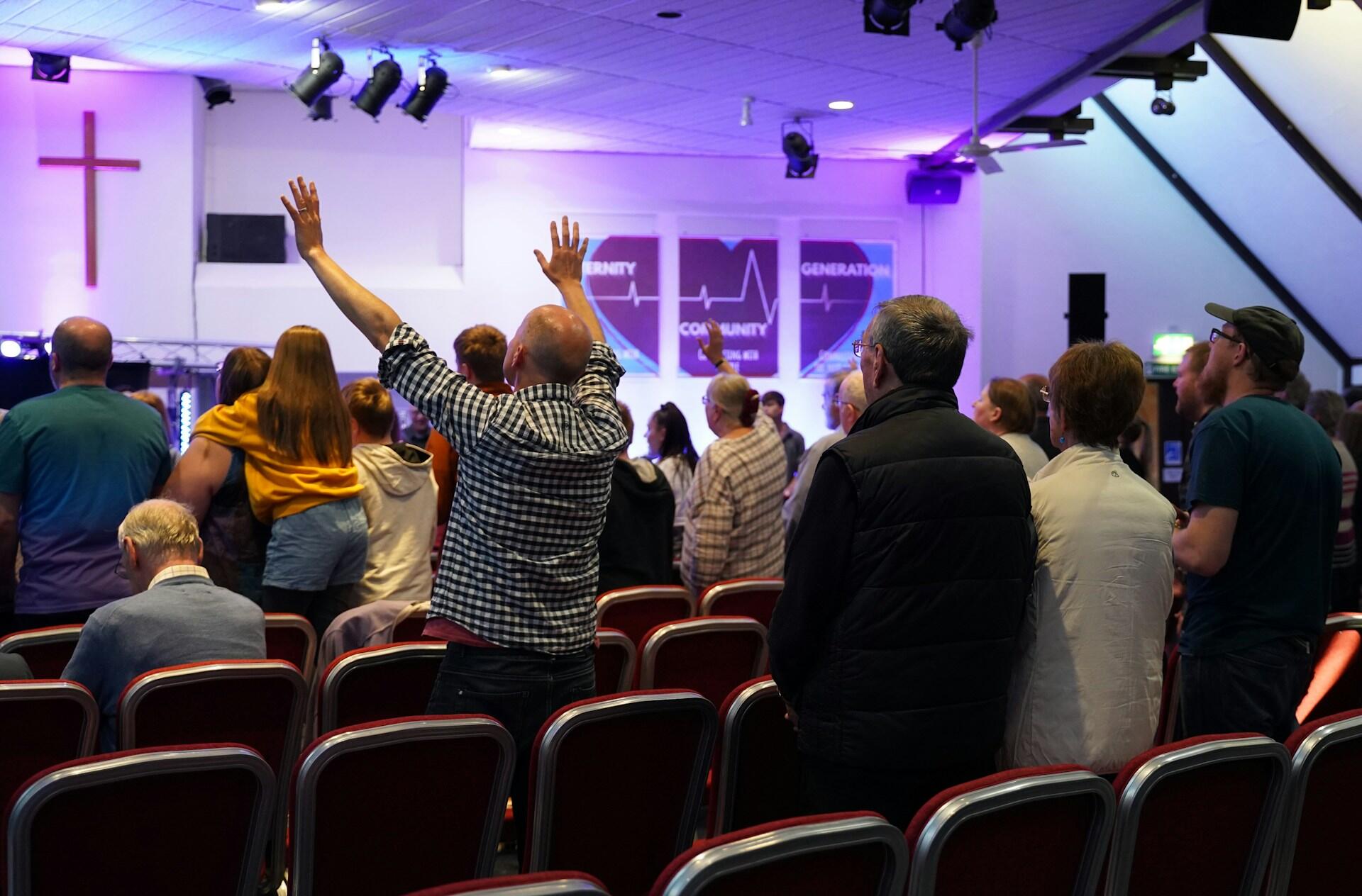 A group of people worshiping in a church, with hands raised, colorful lighting, and a backdrop featuring a cross and screens.