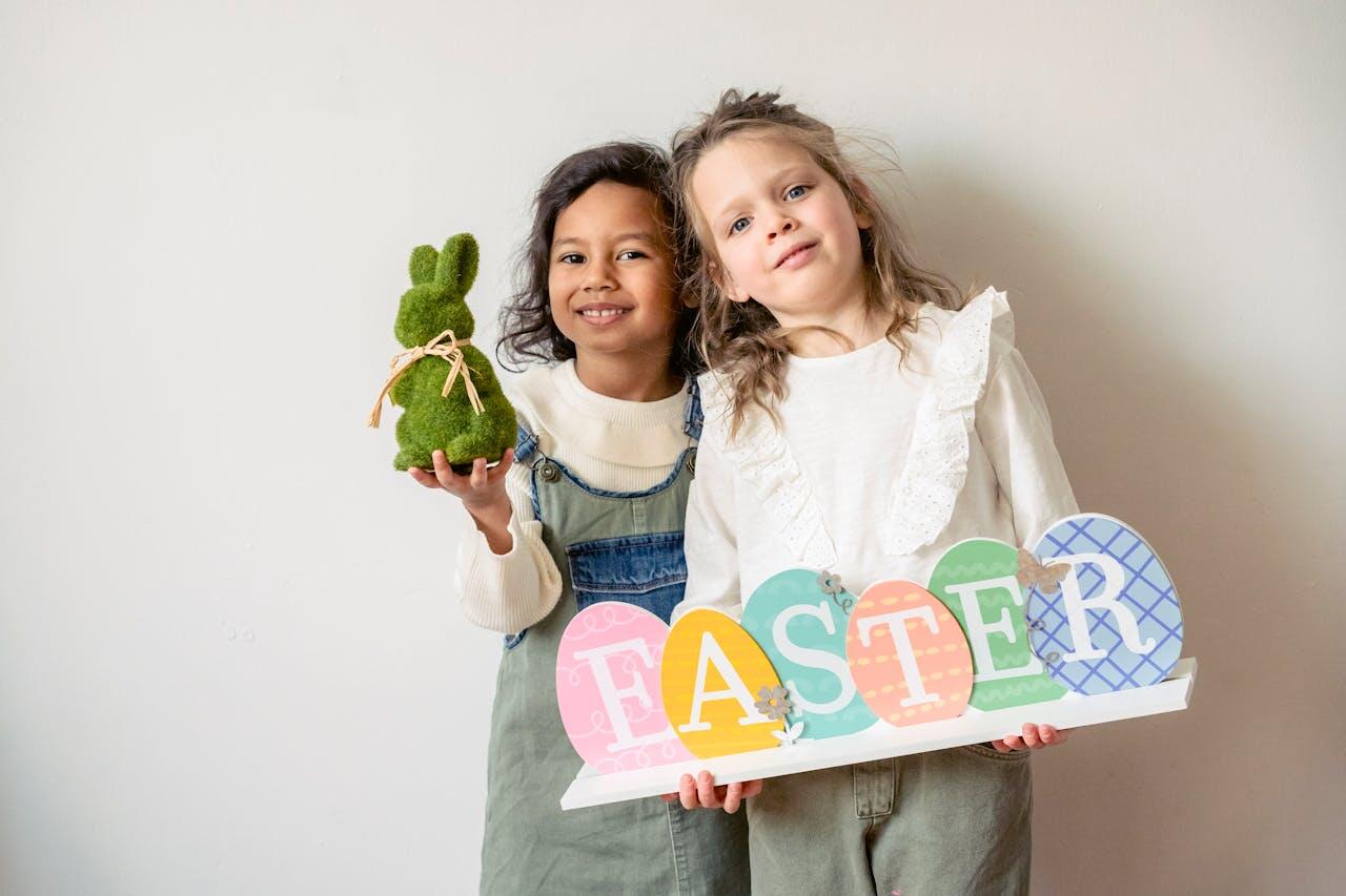 Two children hold a decorative sign that reads "EASTER" and a green bunny figure, set against a simple, light backdrop.