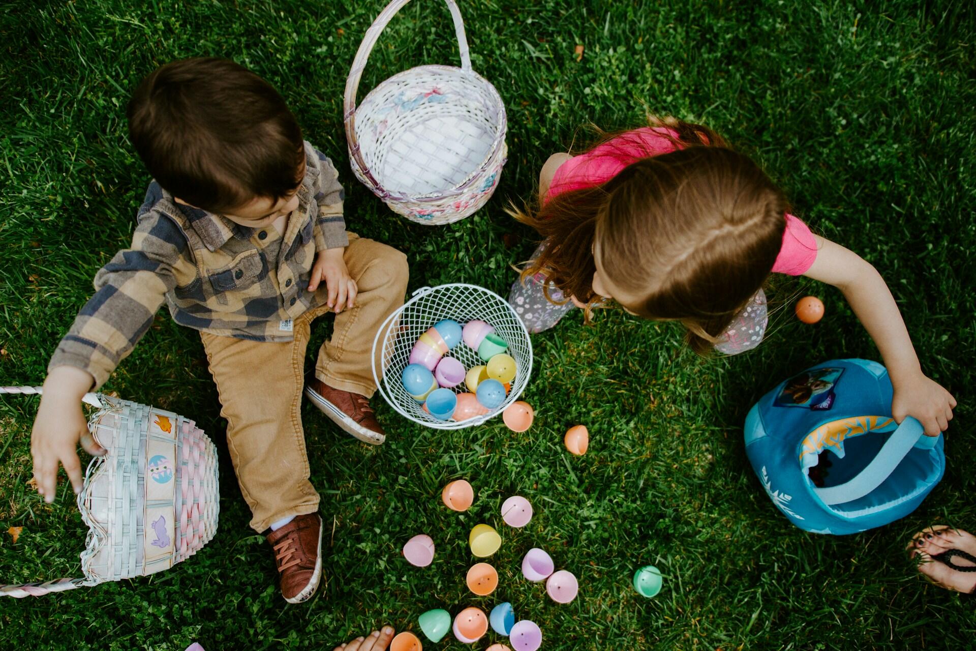 Two children gather colorful plastic Easter eggs on grass, with baskets nearby, one child holding a blue bag.