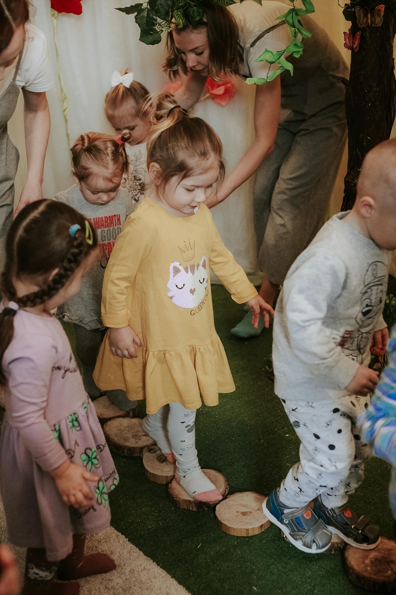 Children engage in a fun activity, balancing on wooden disks on a grassy floor, with adults assisting them. Bright colors and playful attire enhance the scene.