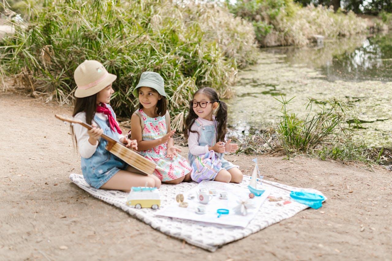 Three children sit on a blanket near a pond, engaged in play with toys and a musical instrument, surrounded by greenery.