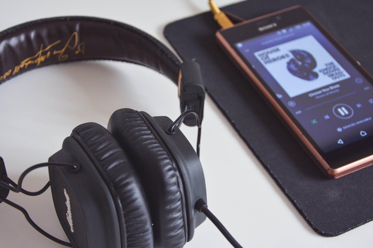A close-up of black headphones next to a smartphone displaying a music streaming app, on a black mouse pad.