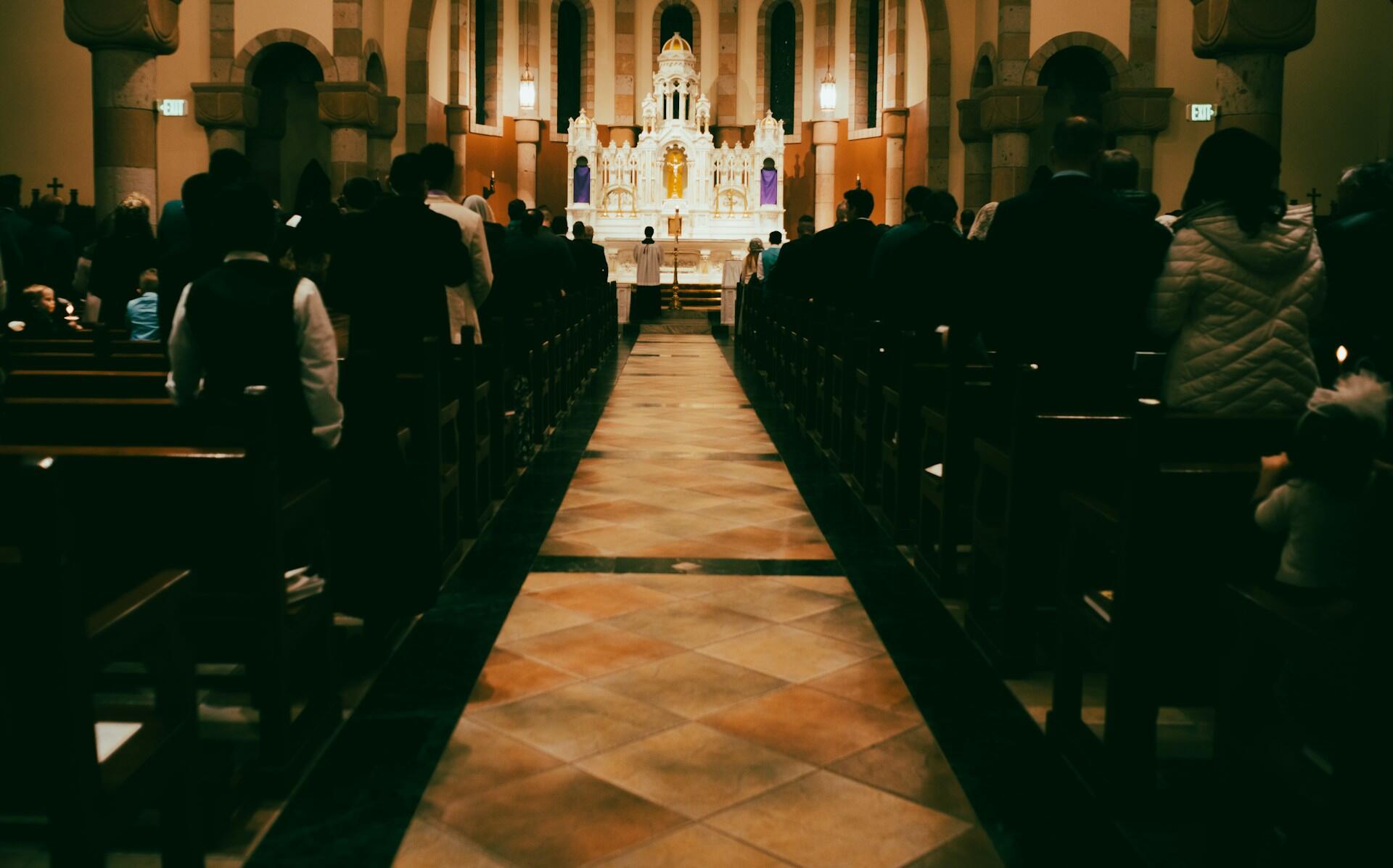 Interior of a church filled with congregants, featuring a decorated altar at the front and wooden pews lining the aisle.