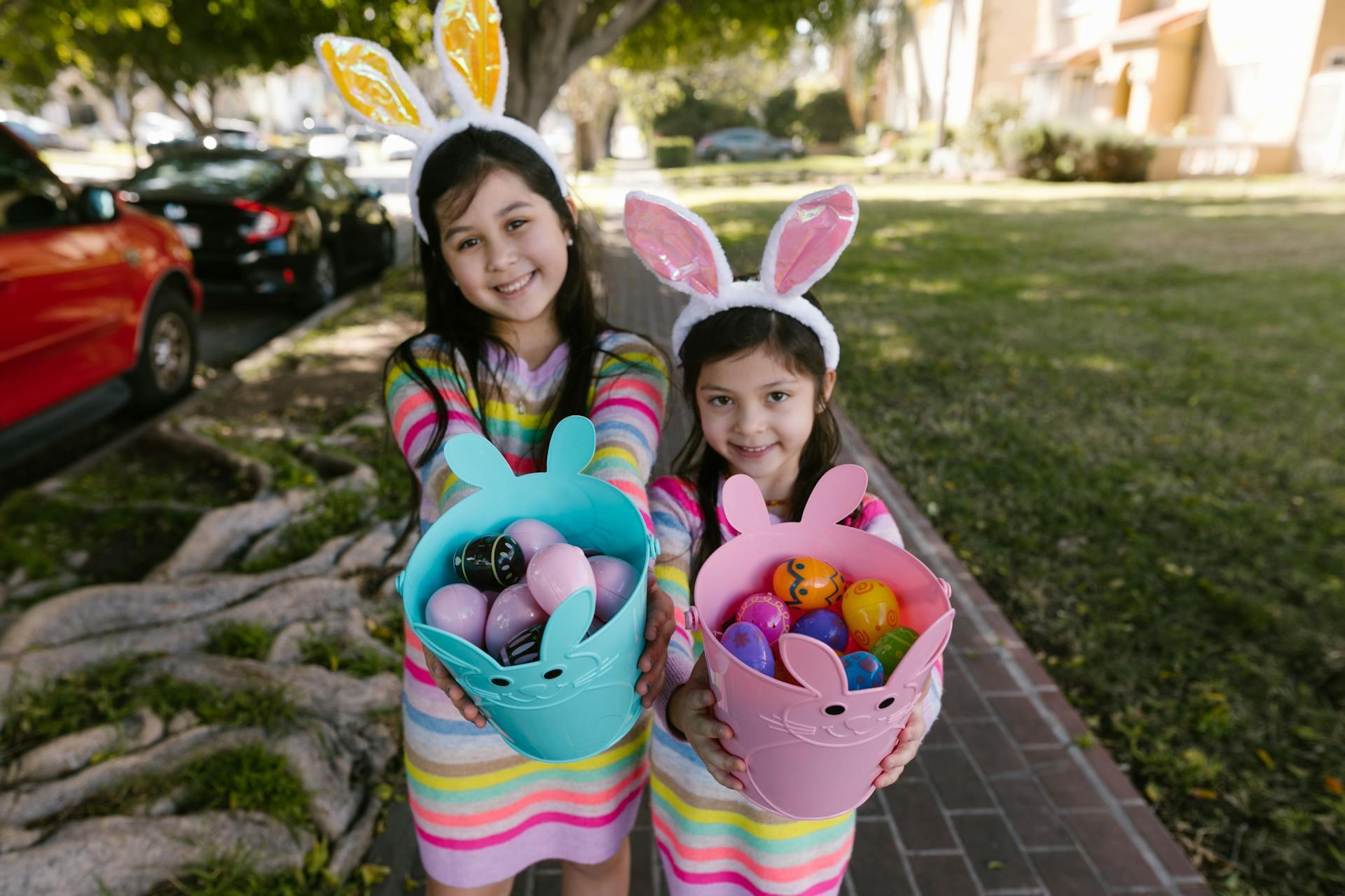 Two children wearing bunny ears hold colorful Easter baskets filled with decorated eggs, standing in a park-like setting.