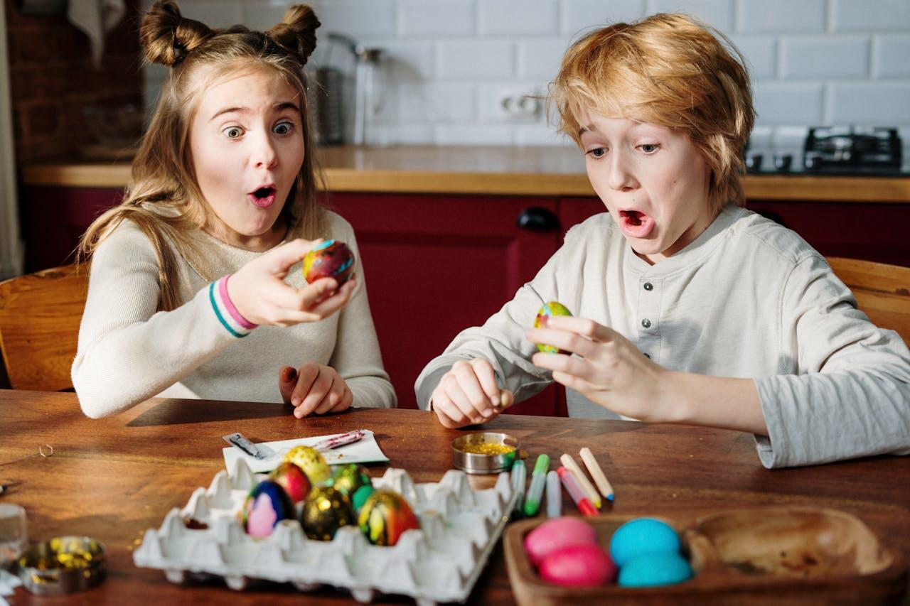 Two children are engaged in painting colorful Easter eggs at a wooden table, surrounded by art supplies and decorated eggs.