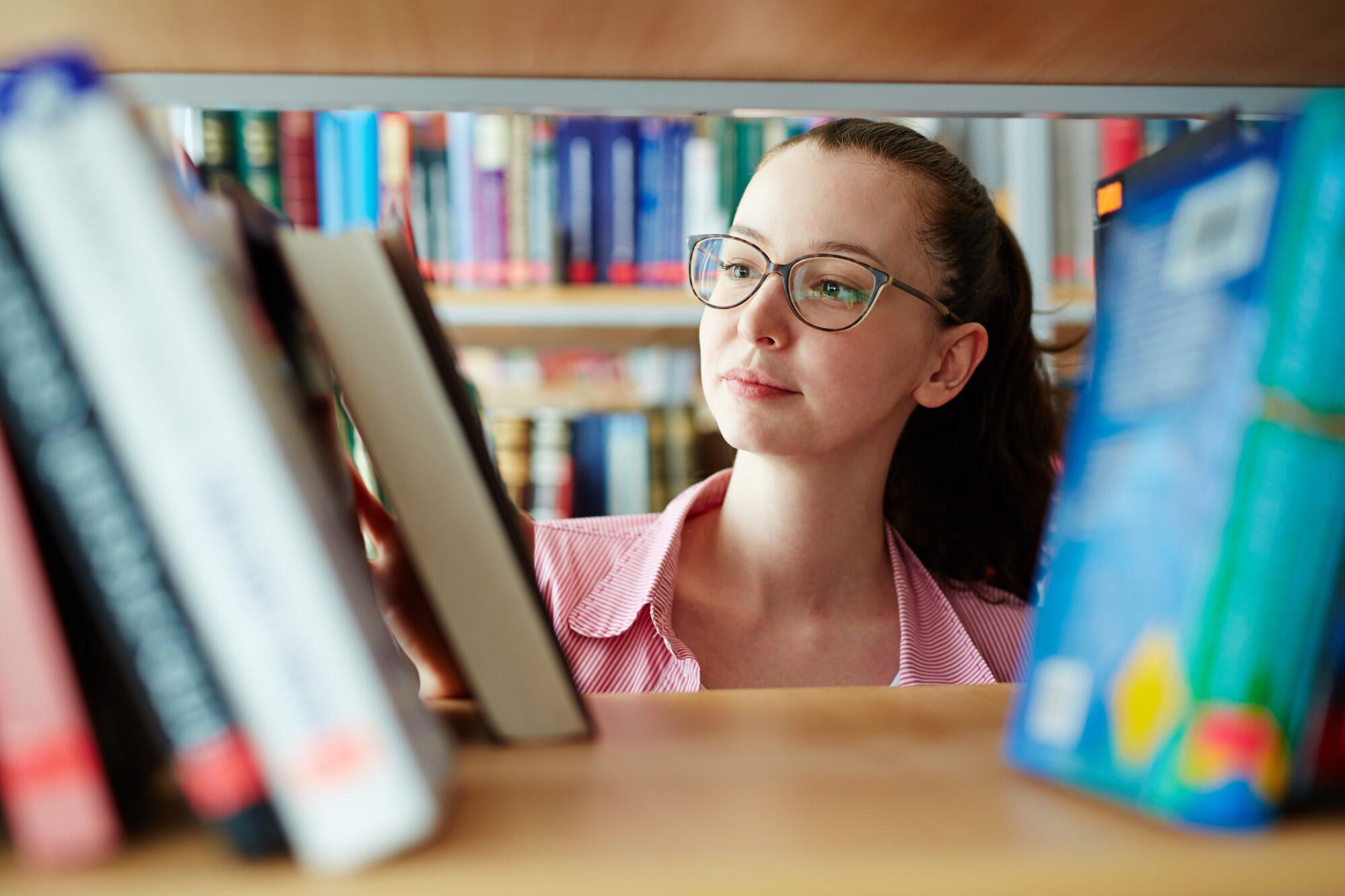 Person with glasses browsing books on a shelf in a library, view through the gap between books.