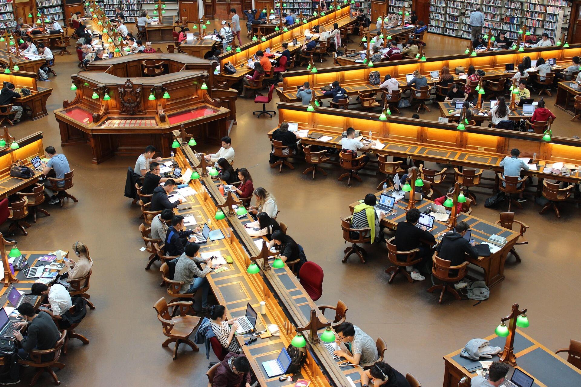 Busy library reading room with many people studying at wooden desks with green lamps and bookshelves along the walls.