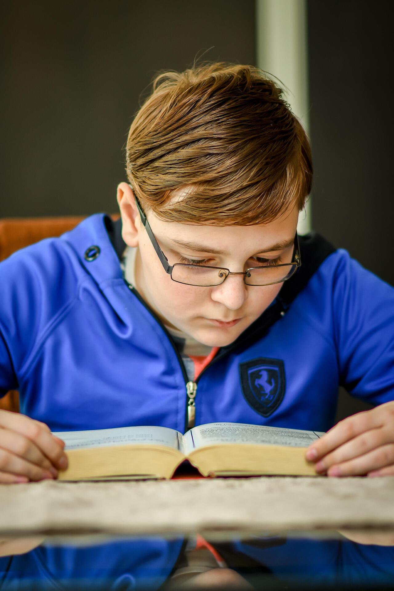 A child wearing a blue jacket is sitting at a table reading an open book.