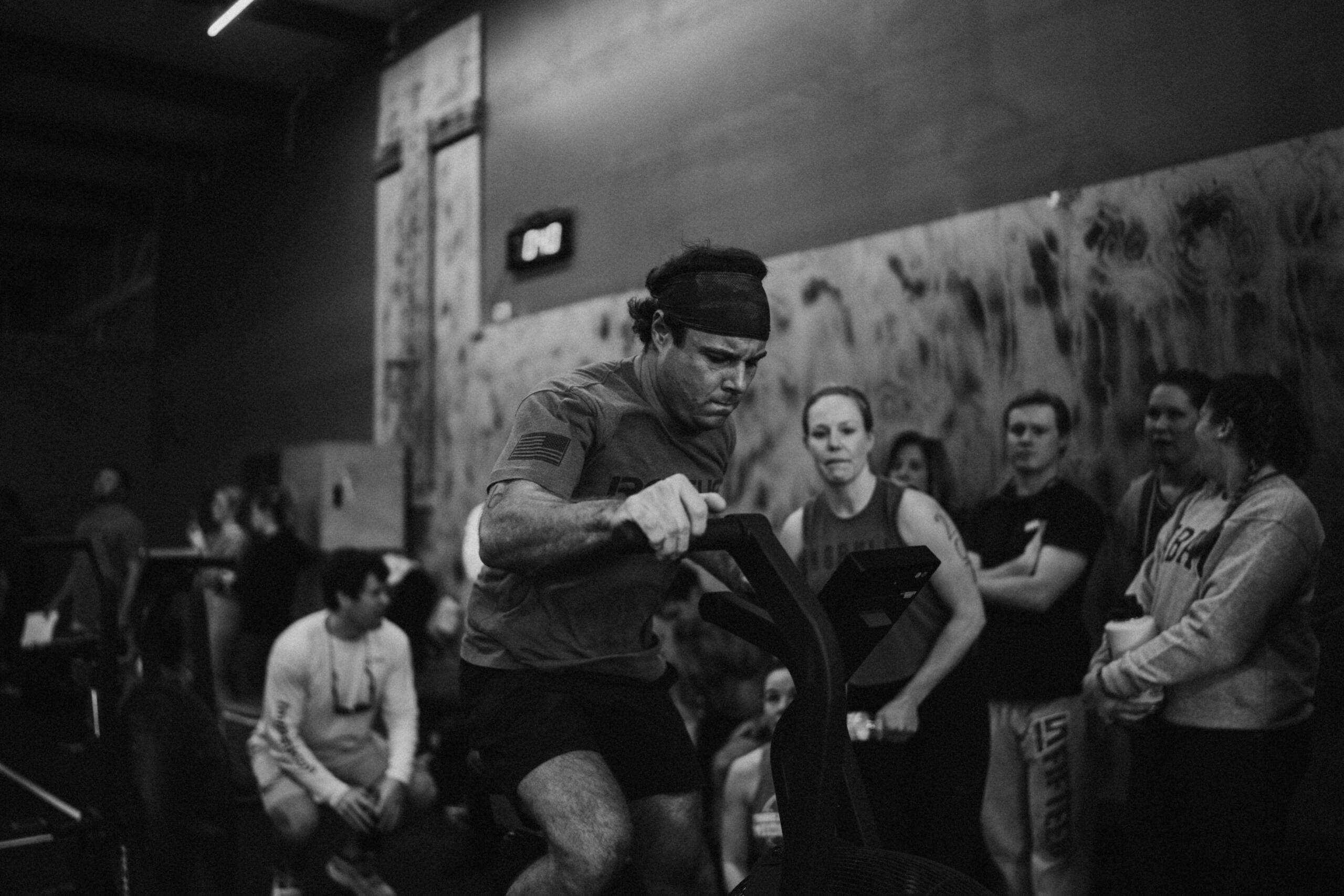 A man using an assault bike with other athletes watching.