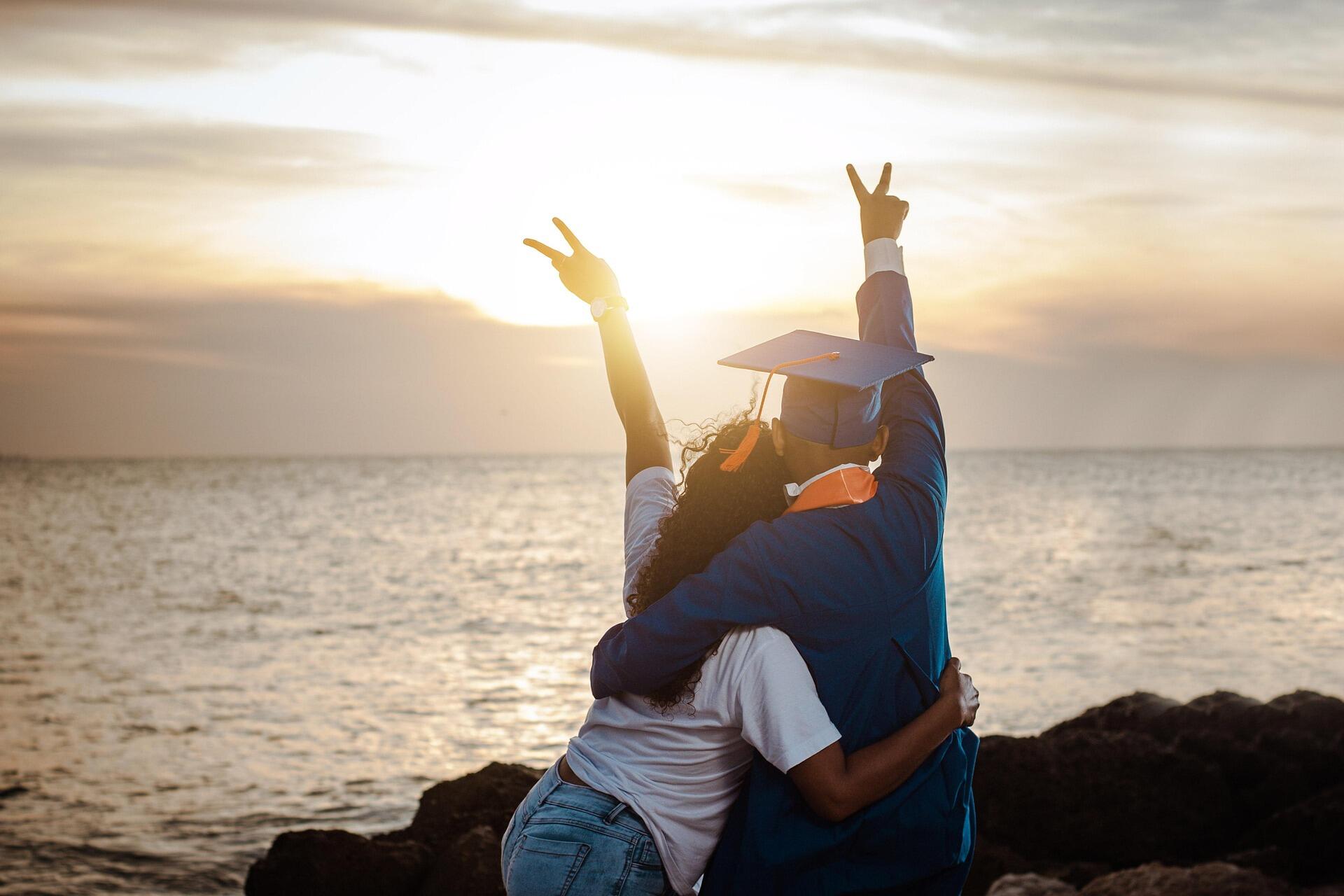 Two people embrace by the ocean at sunset, one wearing a graduation cap and gown, both raising their hands in peace signs.