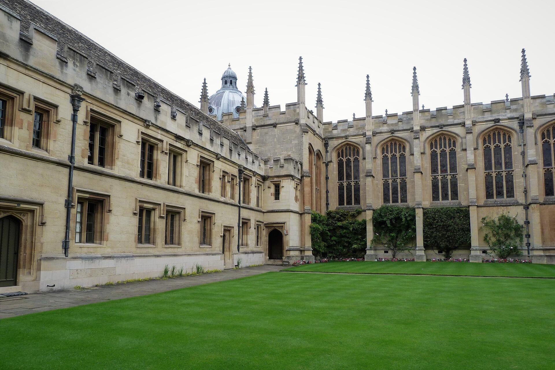 Courtyard of a historic stone building with large arched windows, gothic spires, and a well-manicured green lawn.