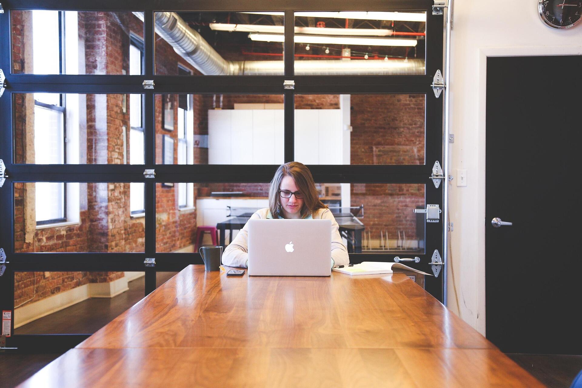 Person working on a laptop at a wooden table in a modern office with exposed brick walls and large glass garage-style doors.