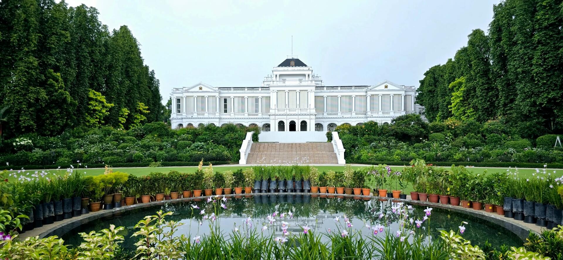 White colonial-style mansion with symmetrical windows, surrounded by lush gardens, large green trees, and a reflective circular pond with flowers in pots.