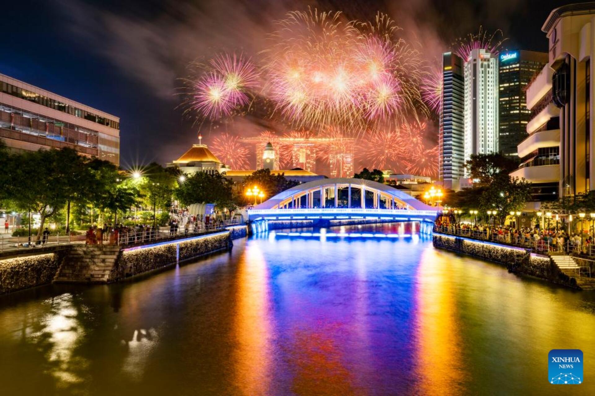 Nighttime cityscape with a lit bridge over a river, colorful fireworks illuminating the sky and reflections in the water.