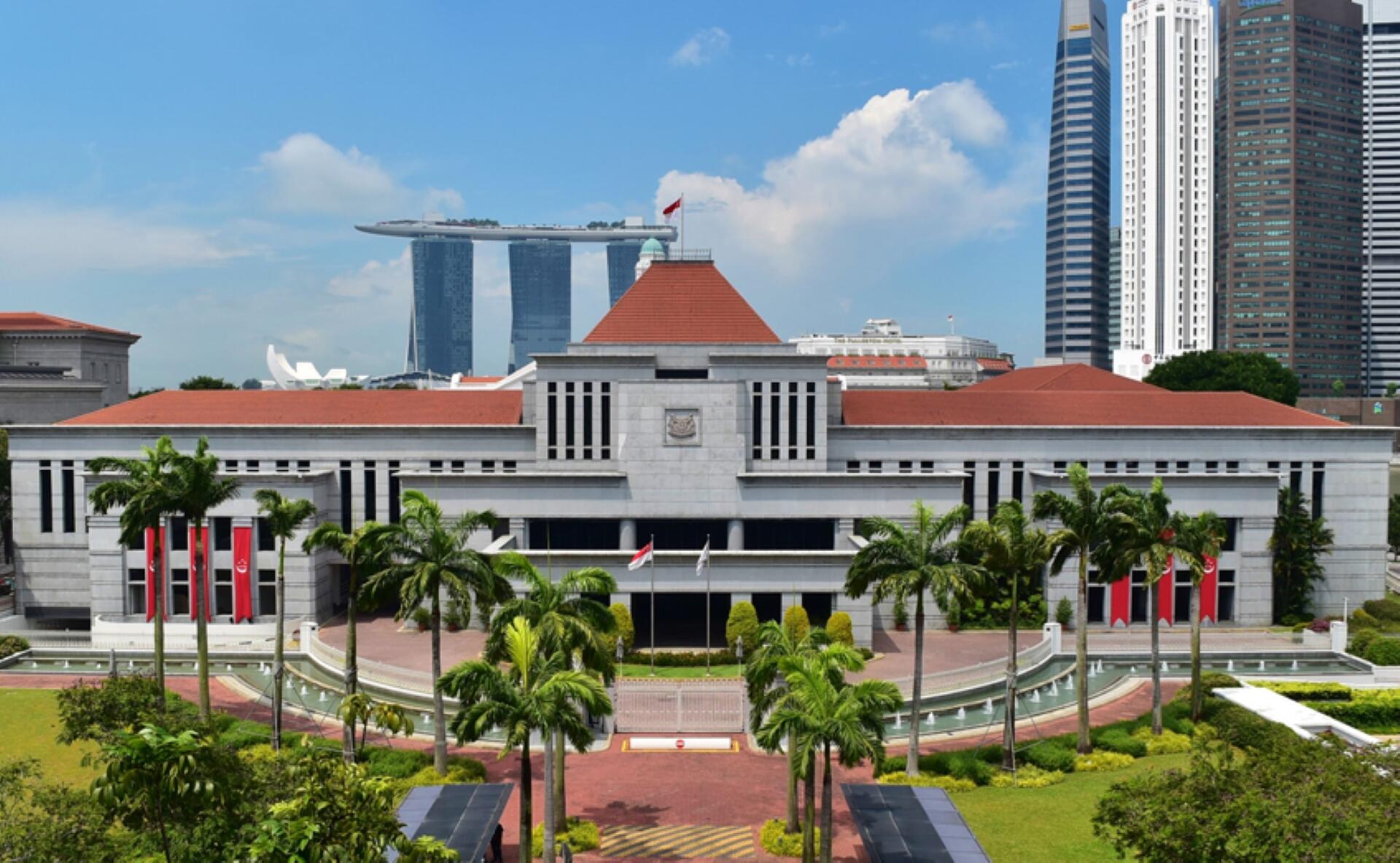 Parliament House of Singapore with palm trees, Singapore flags, and Marina Bay Sands in the background under a blue sky.