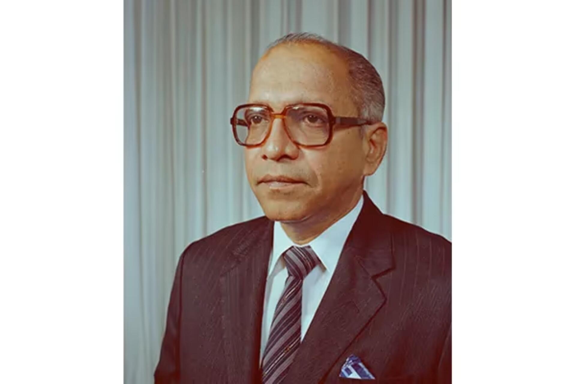 Portrait of Deven Nair in a dark pinstripe suit, white shirt, striped tie, and blue pocket square, standing against a light curtain background.