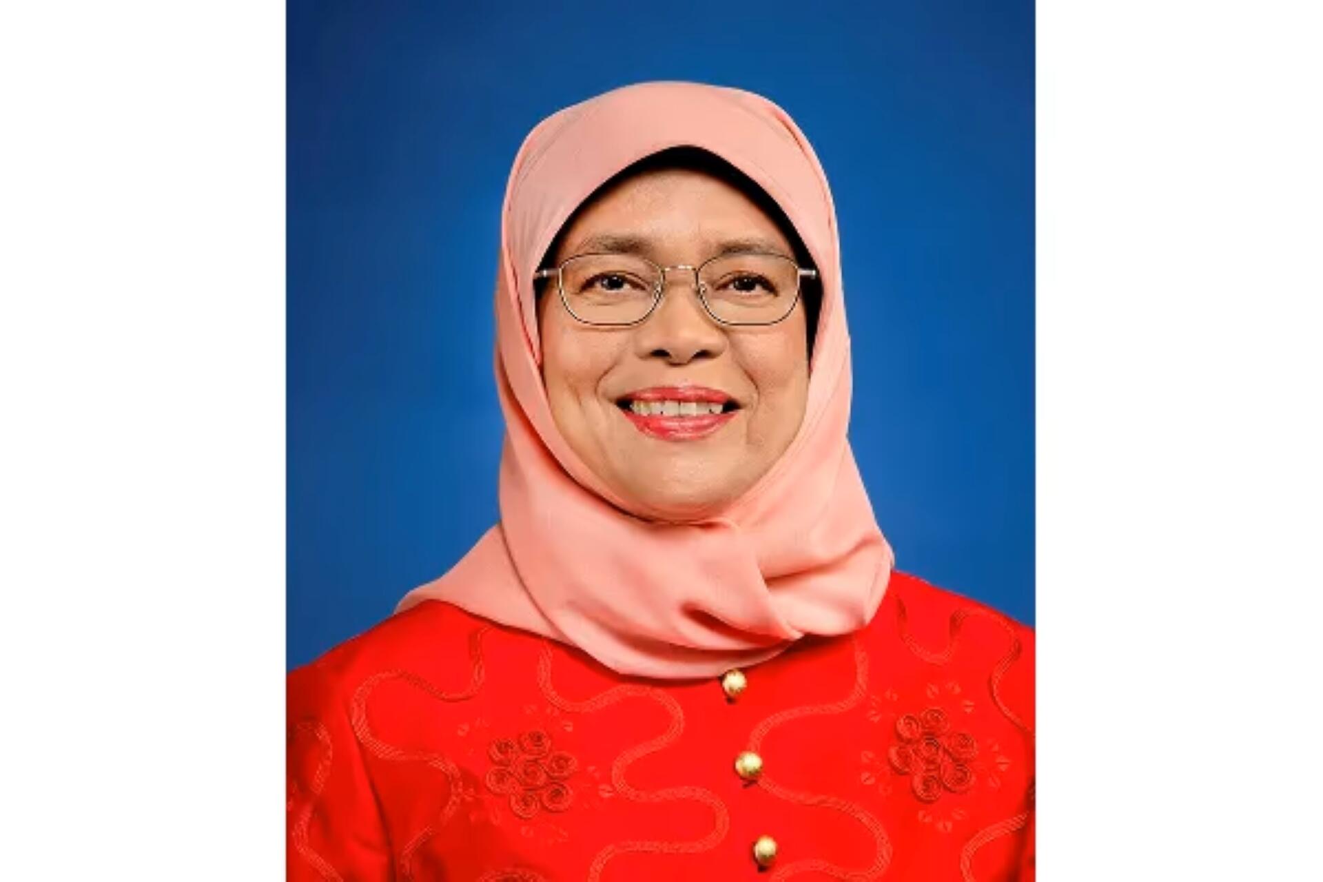 Halimah Yacob wearing glasses and a light pink hijab over a bright red blouse smiles gently against a solid blue background. Her expression is warm and welcoming.