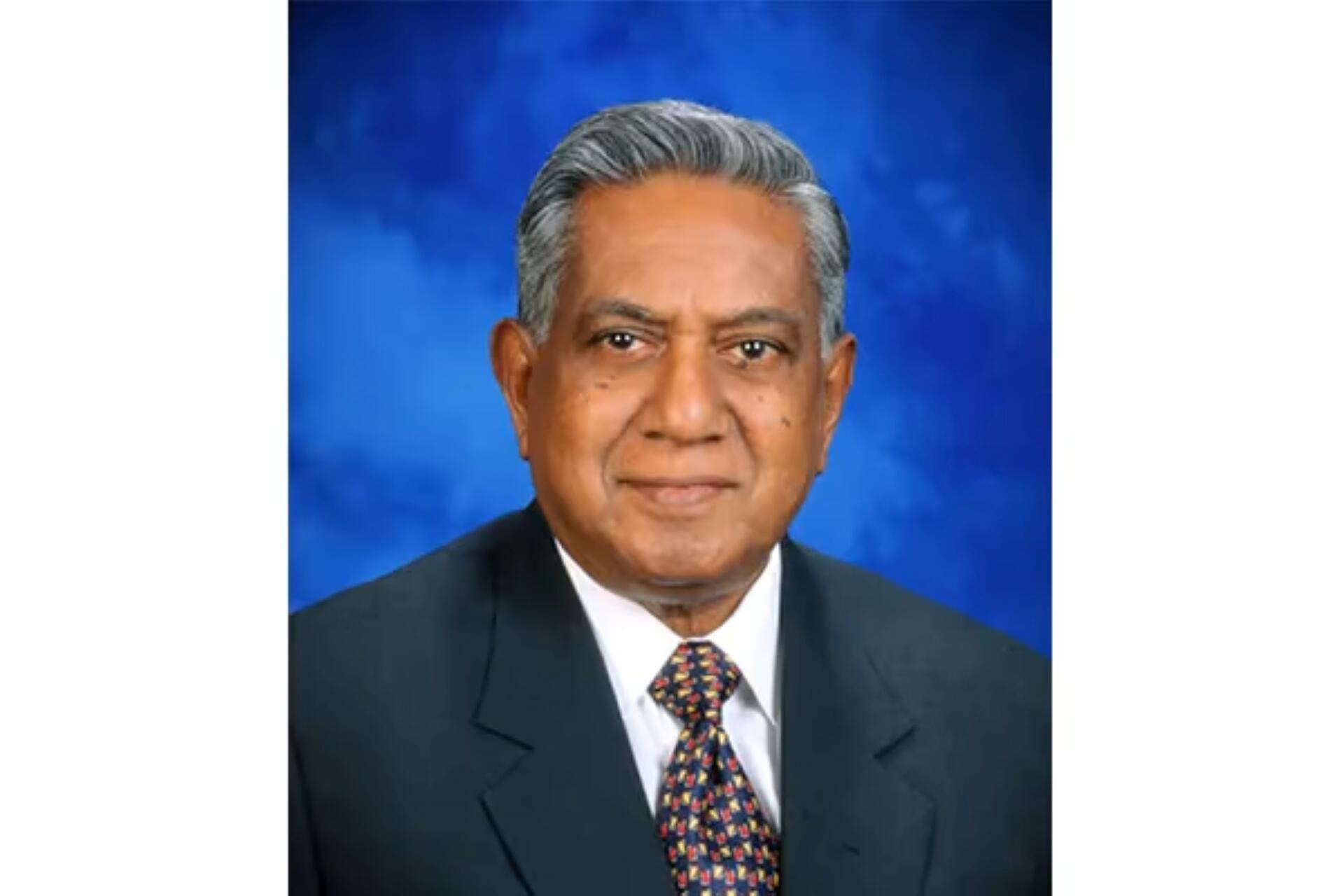 Portrait of S R Nathan in a dark suit, white shirt, and colorful tie against a blue studio background.