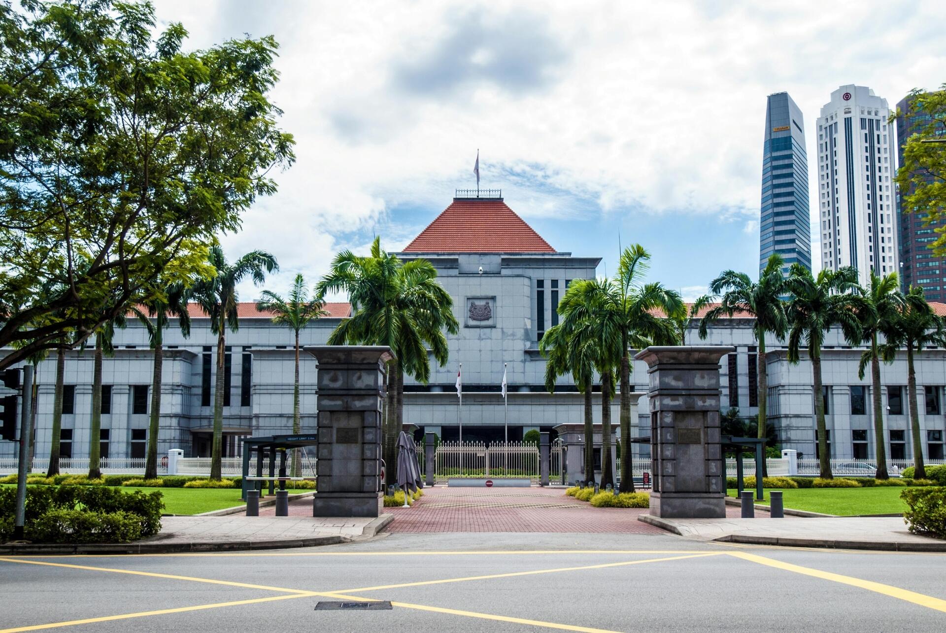 View of a grand government building with a red-tiled roof, palm trees, and skyscrapers in the background on a partly cloudy day.