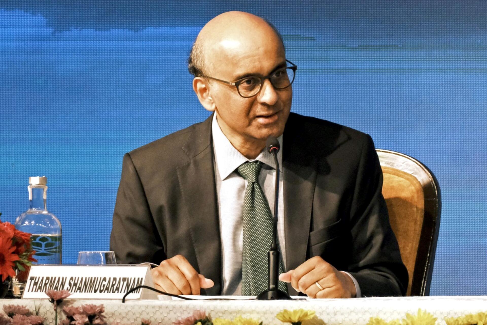 Tharman Shanmugaratnam in a suit speaks into a microphone at a conference. He's seated at a table with a nameplate and flowers.
