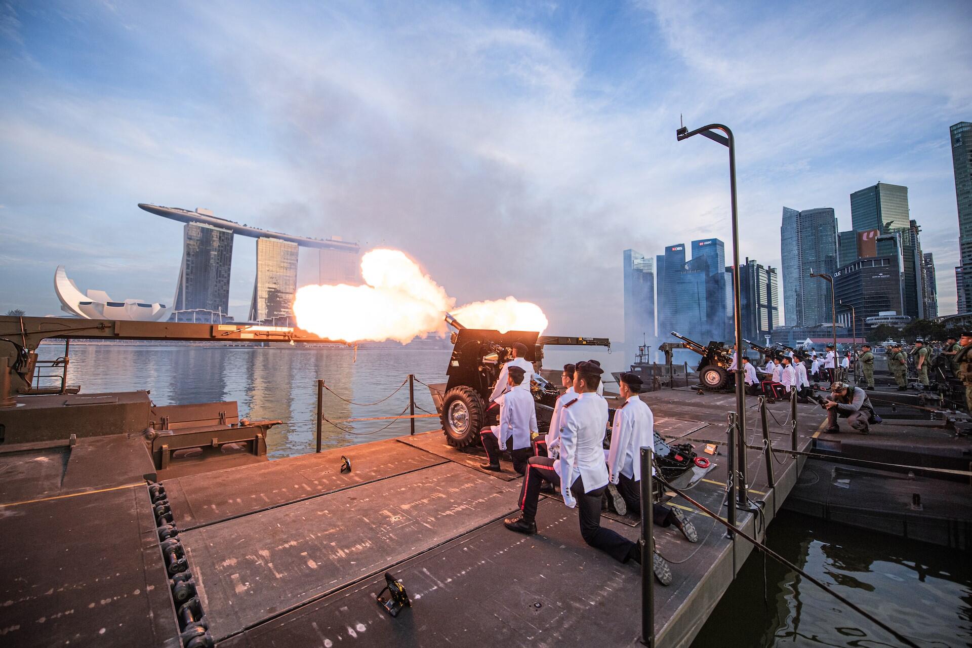 Several individuals in white officer uniforms gather on a dock next to a cannon, with a scenic waterfront view in the background.