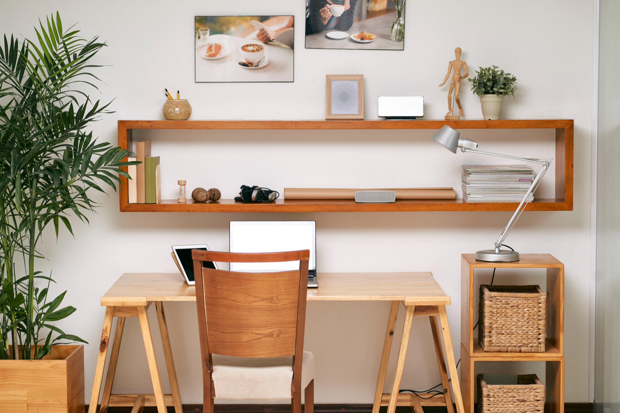Home office setup with wooden desk, chair, laptop, tablet, floating shelf with books, lamp, plant, and framed coffee-themed pictures on the wall.