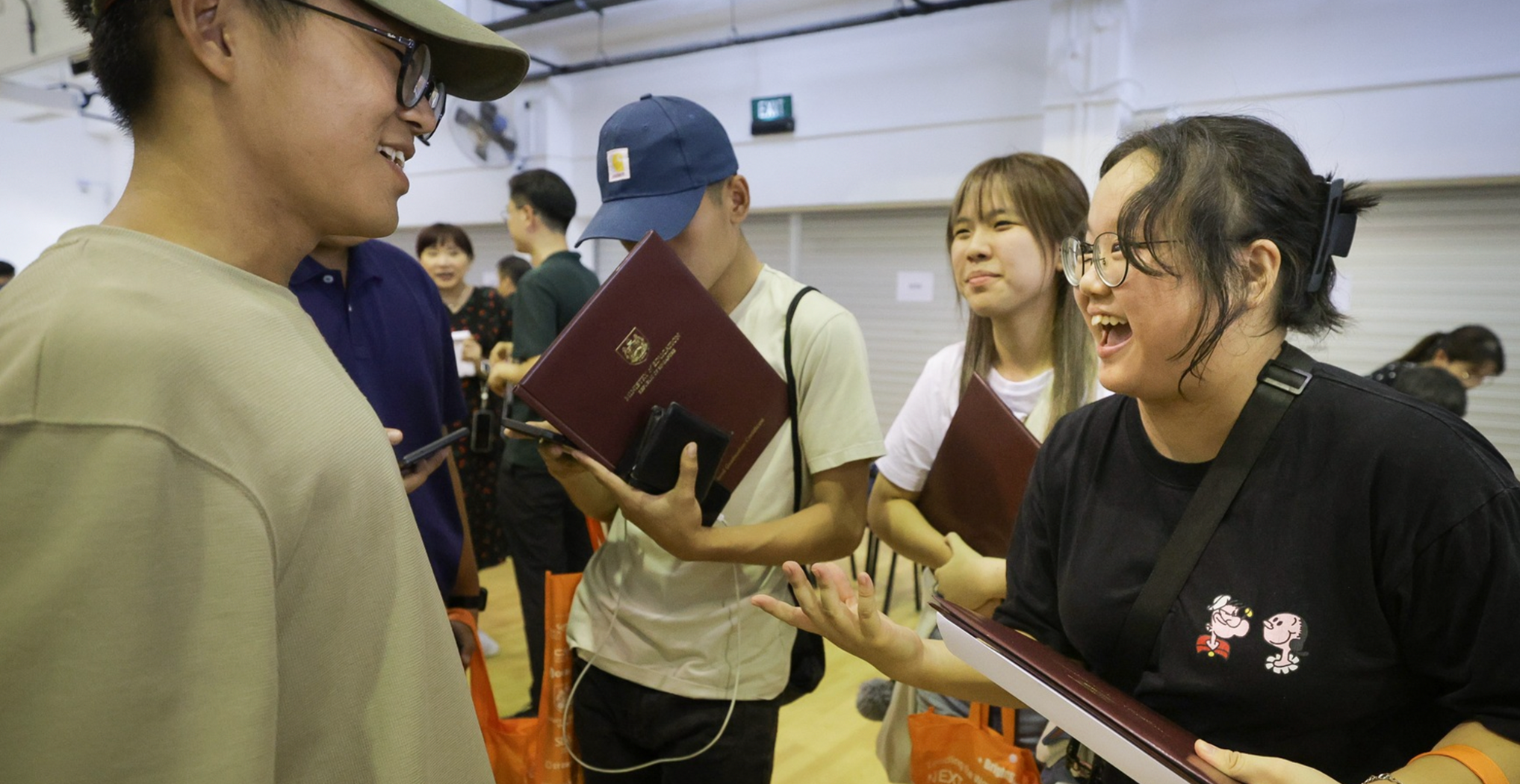A group of young adults celebrates joyfully indoors. One holds a diploma. Their expressions are happy and excited, creating a lively and festive mood.
