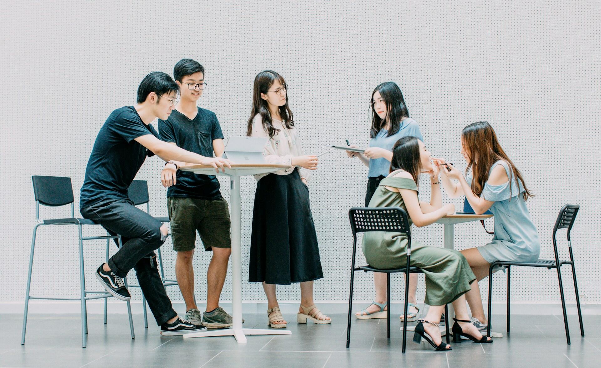 Six young adults are engaged in a casual discussion in a modern, minimalist space. Three are seated at tables while three stand nearby, all appearing focused.