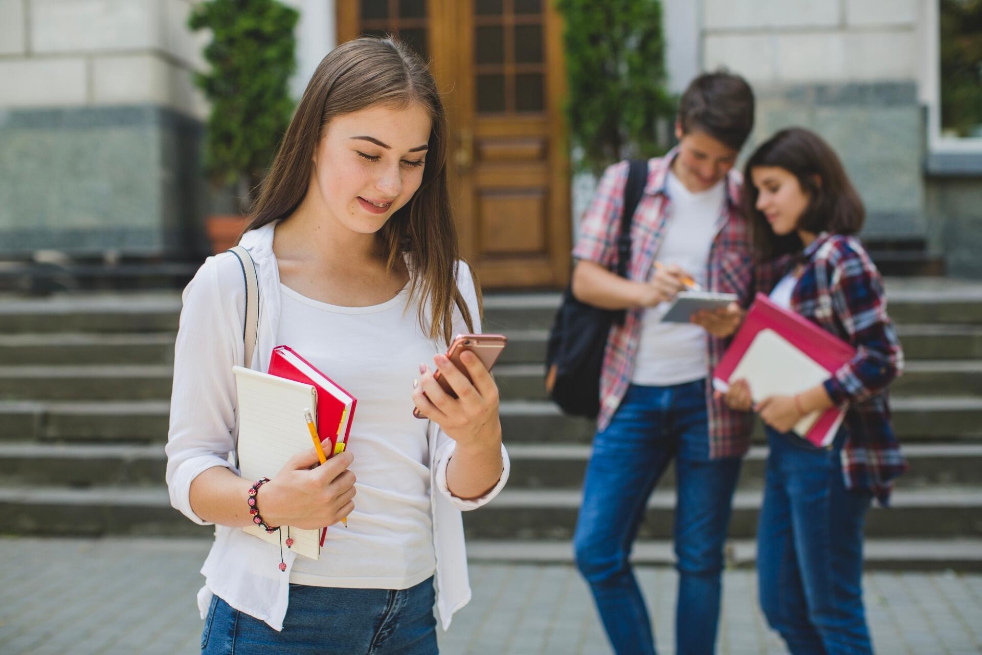 Student holding notebooks and phone with two other students standing and writing in the background outside a building.