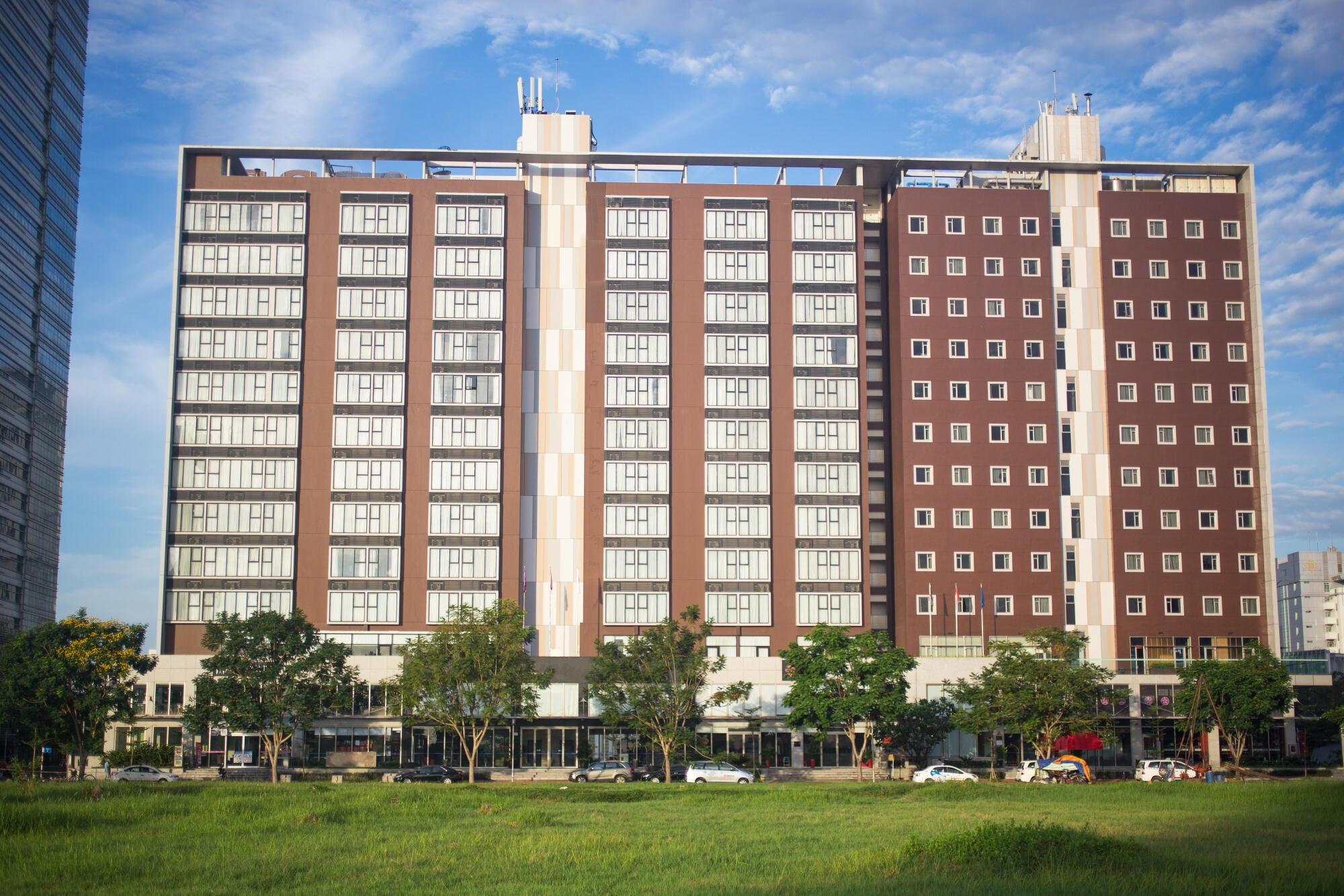 Modern multi-story building with symmetrical windows, trees lining the front, parked cars, and a clear blue sky background.