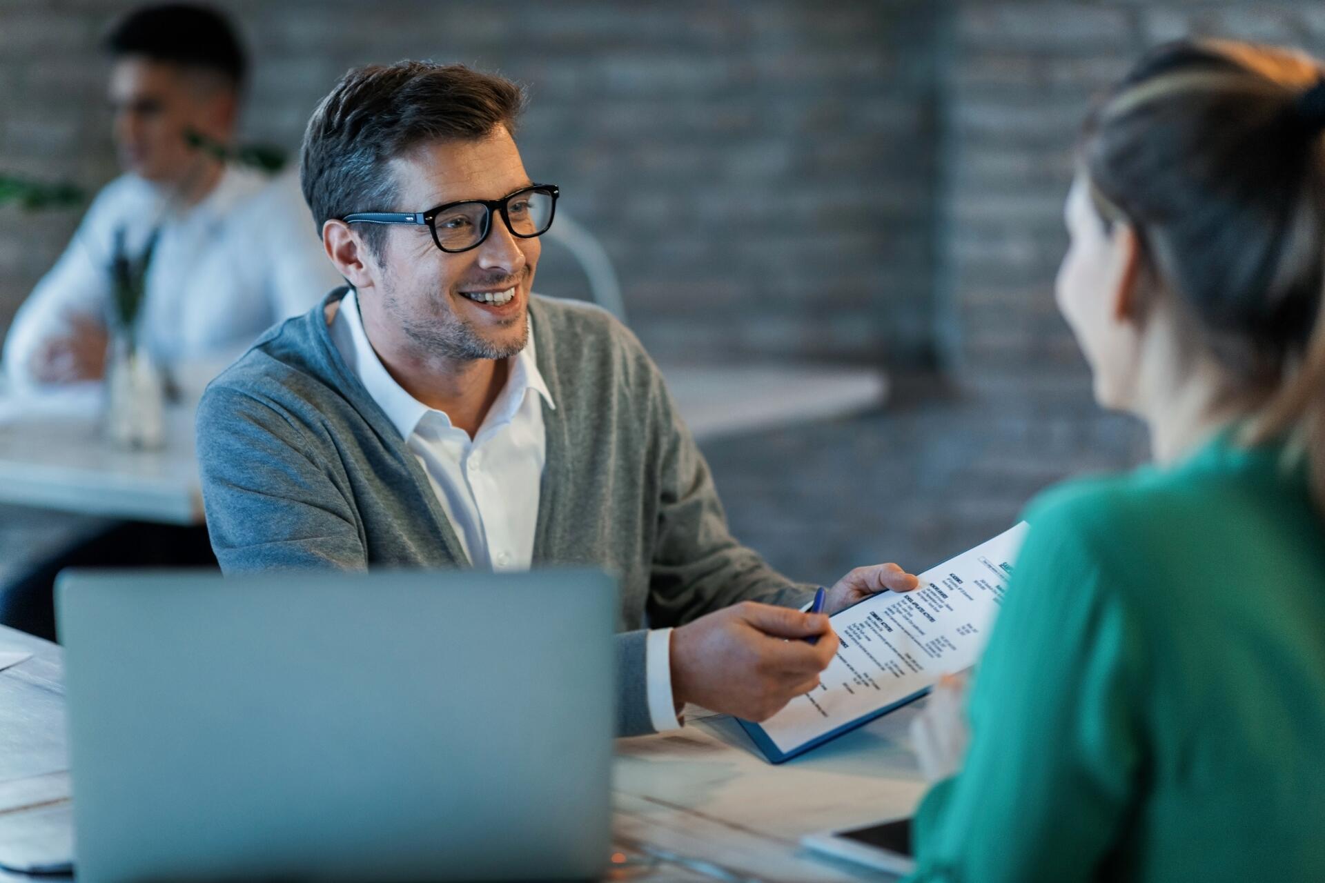 Person in glasses discussing a resume or document with another person in a professional setting, with a laptop on the table.