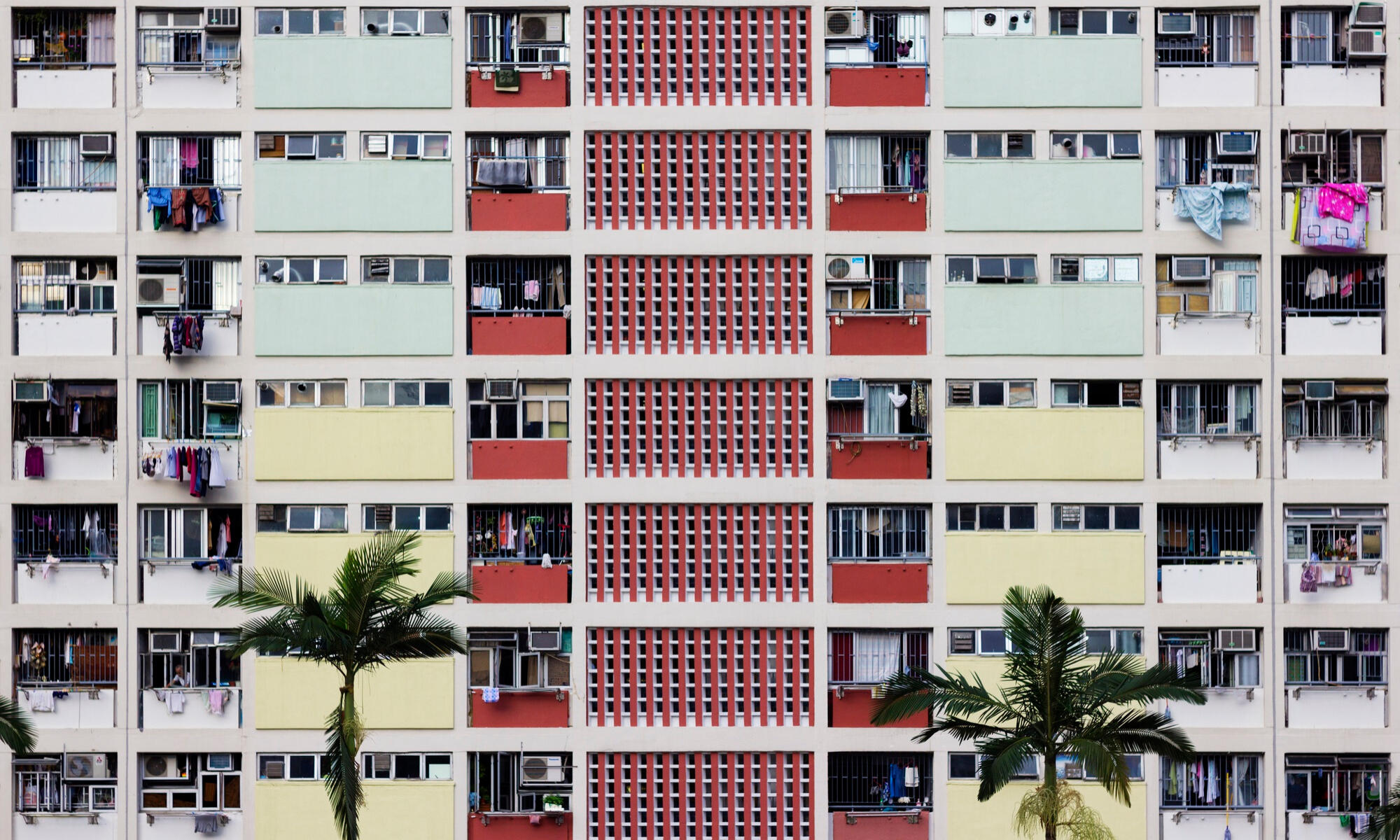Exterior view of a multistory apartment building with colorful panels, barred windows, laundry hanging, and two palm trees in front.