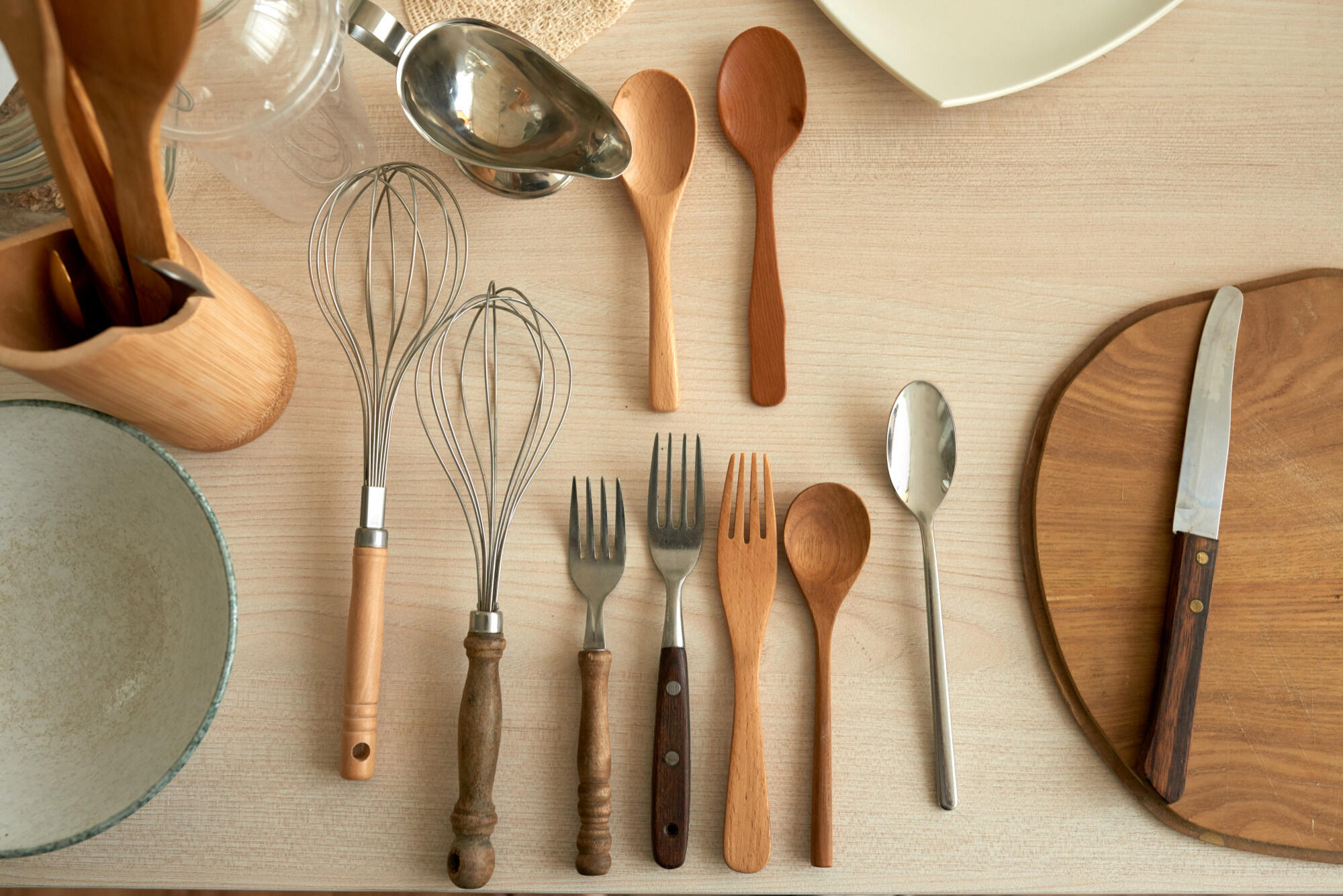 Various wooden and metal kitchen utensils, including whisks, spoons, forks, a knife on a wooden cutting board, and a bowl on a light wood table.