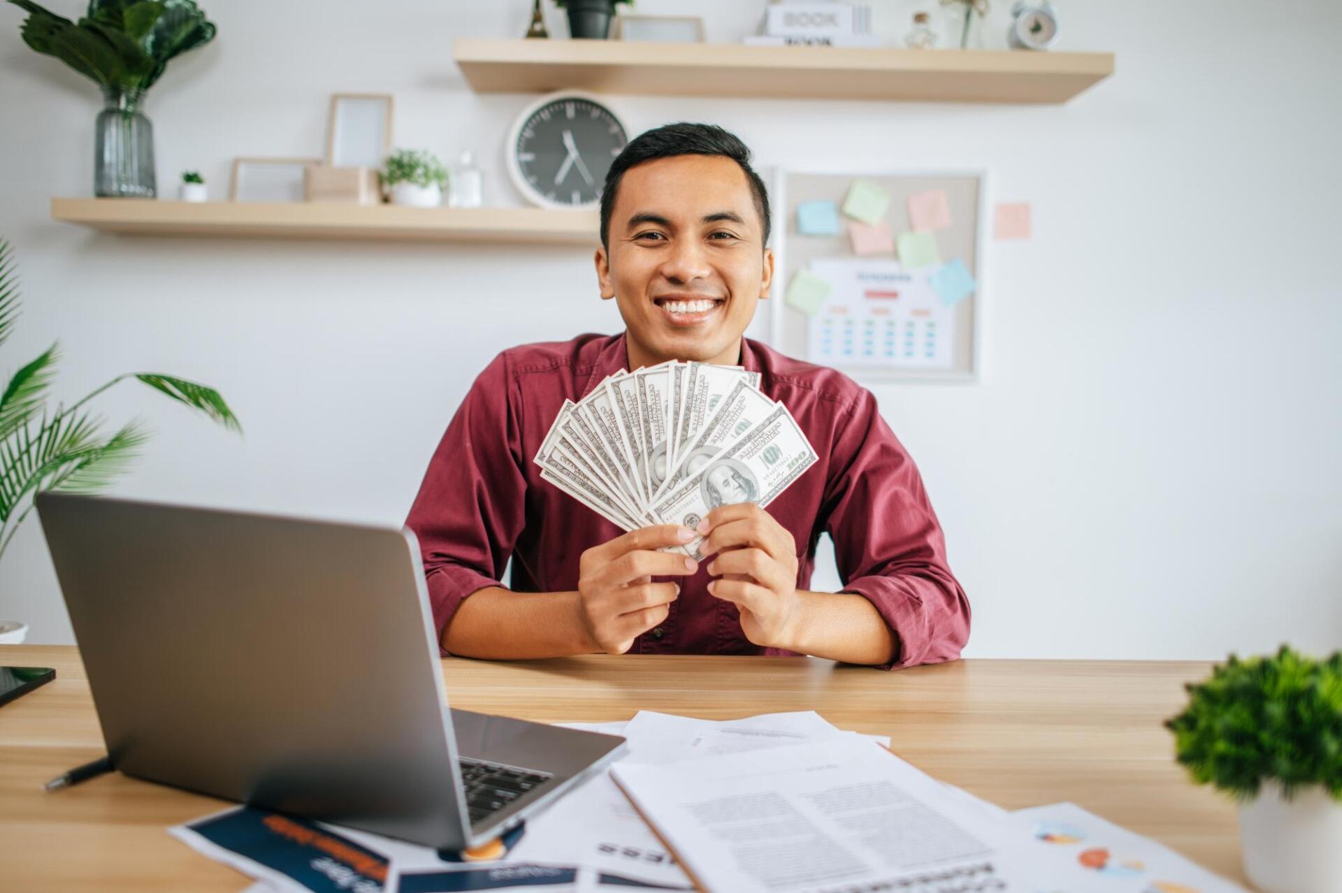 Person seated at desk holding fanned US dollar bills, with laptop and documents on the table in a modern office setting.